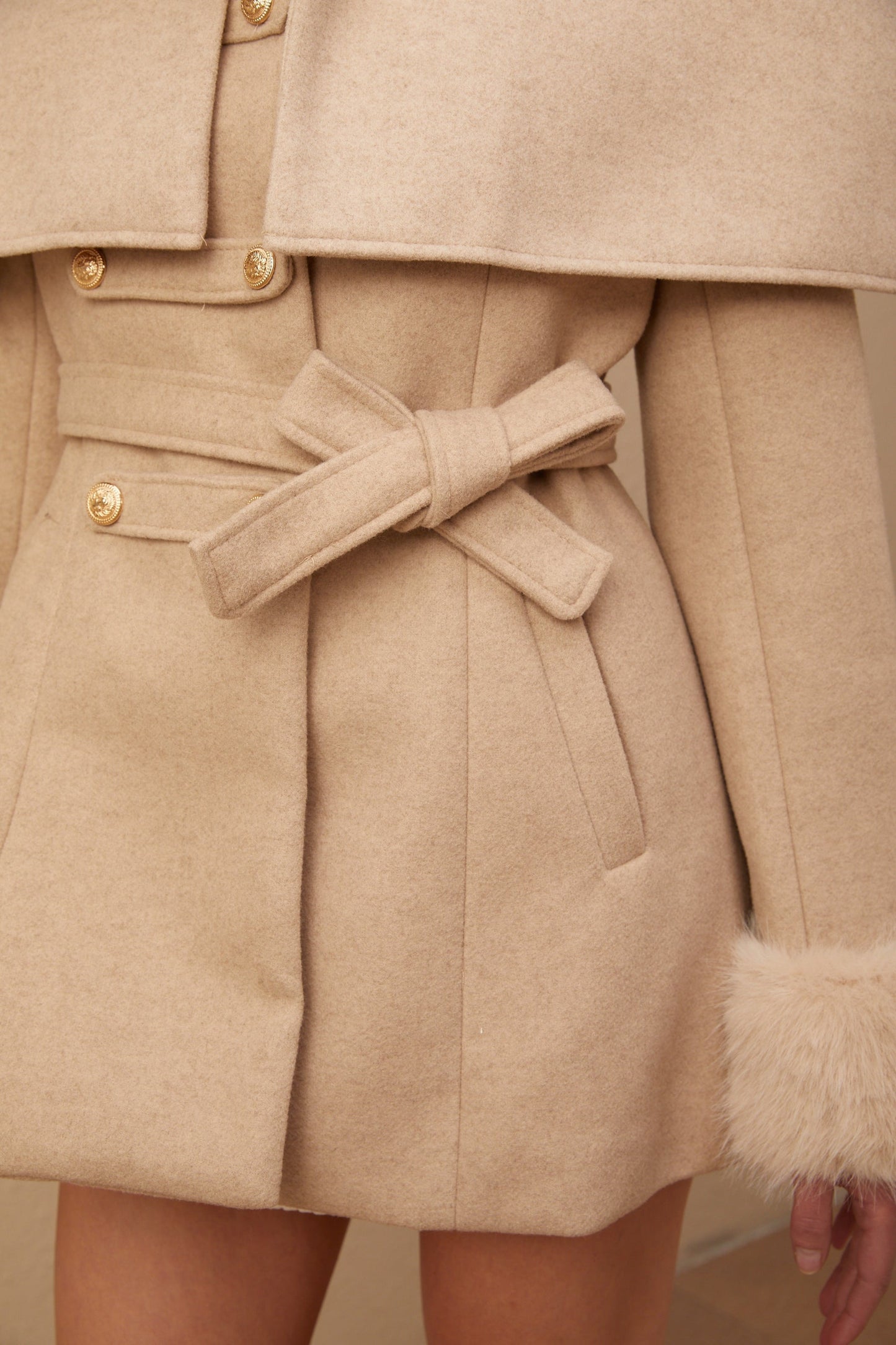 A woman wearing a long black faux fur coat with a detachable cape collar standing against a plain background