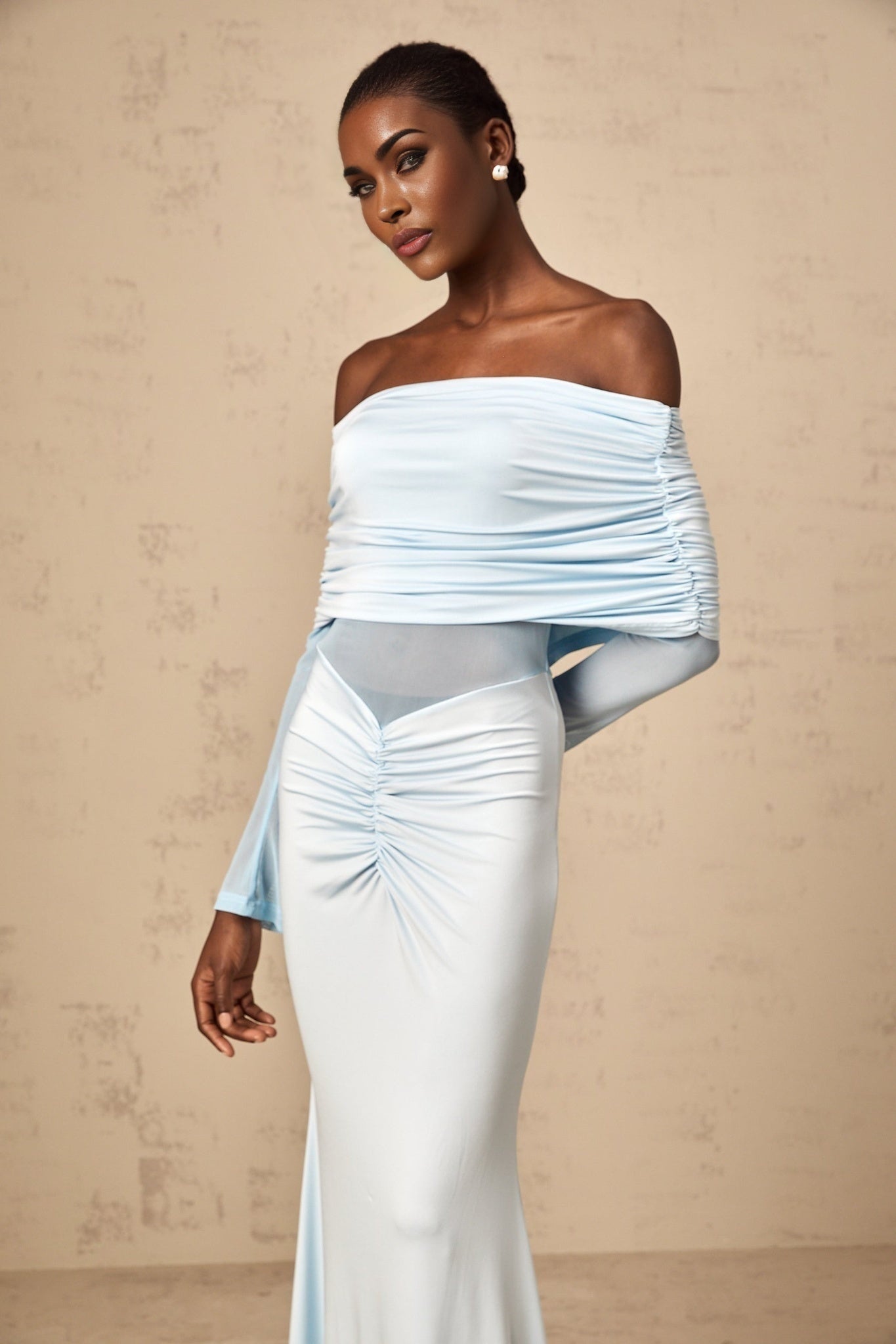 A woman wearing a long blue offshoulder dress with ruched detailing and a flowing maxi skirt