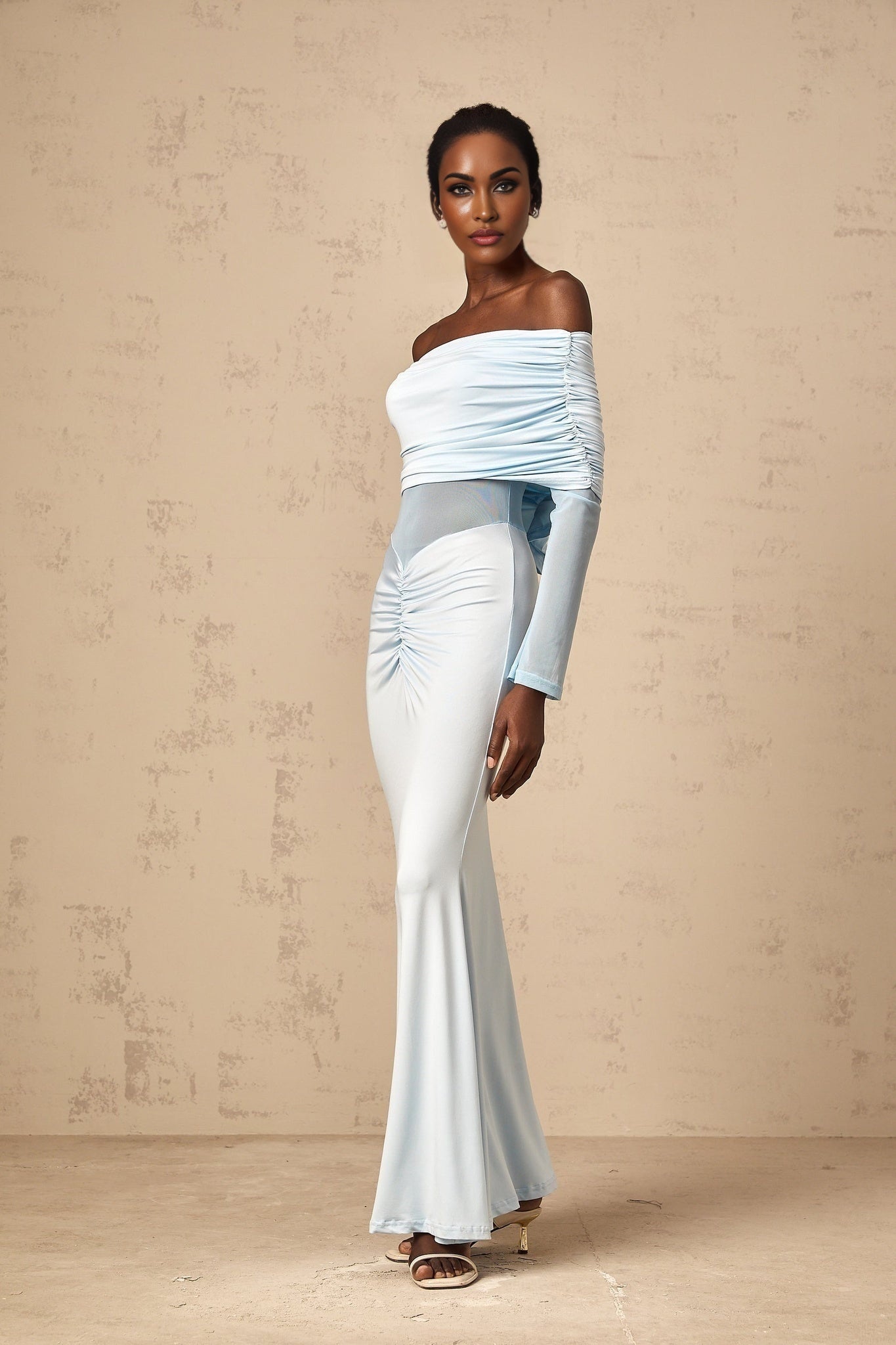 A woman wearing a long blue offshoulder dress with ruched detailing standing against a plain background