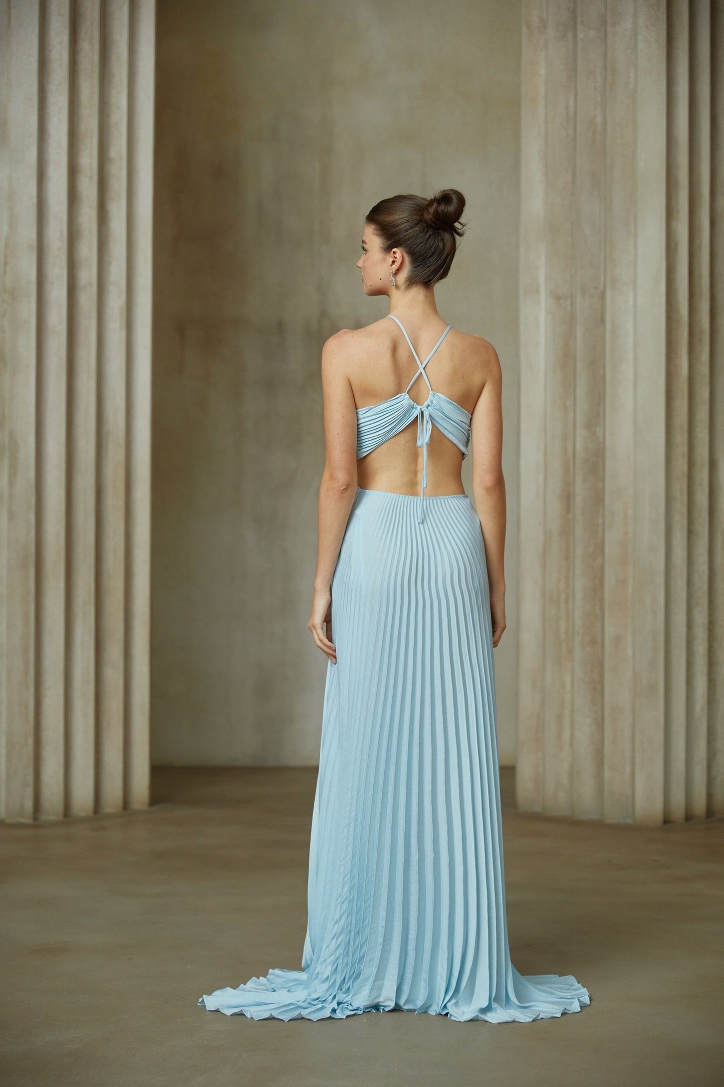 A woman wearing a long blue pleated maxi dress with a cutout detail on the waist standing against a plain background