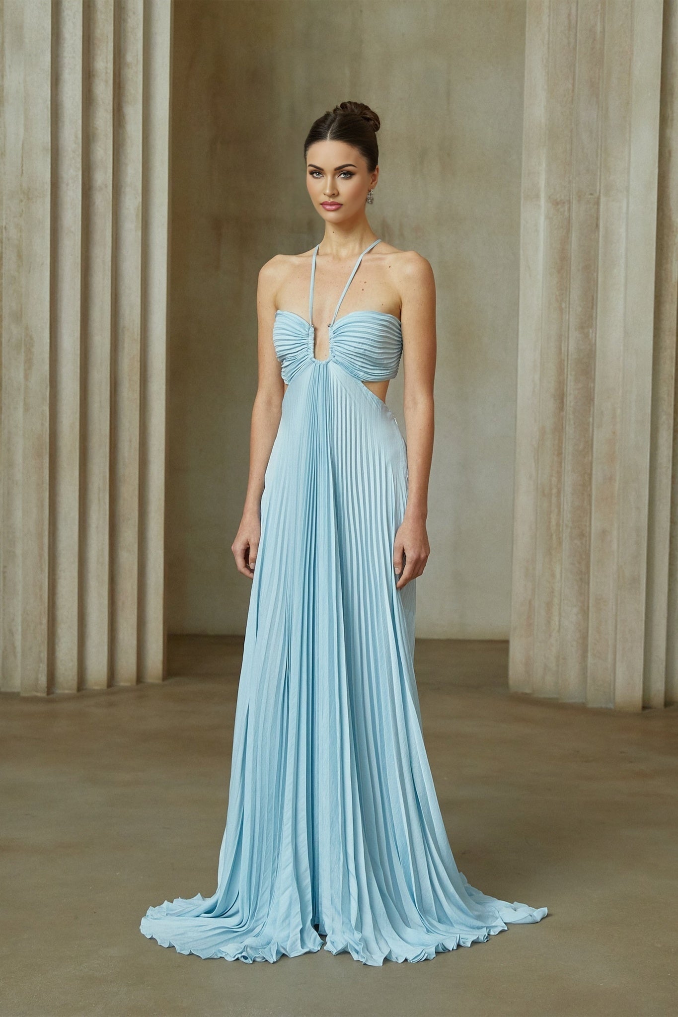 A woman wearing a long blue pleated maxi dress with a cutout detail standing against a plain background