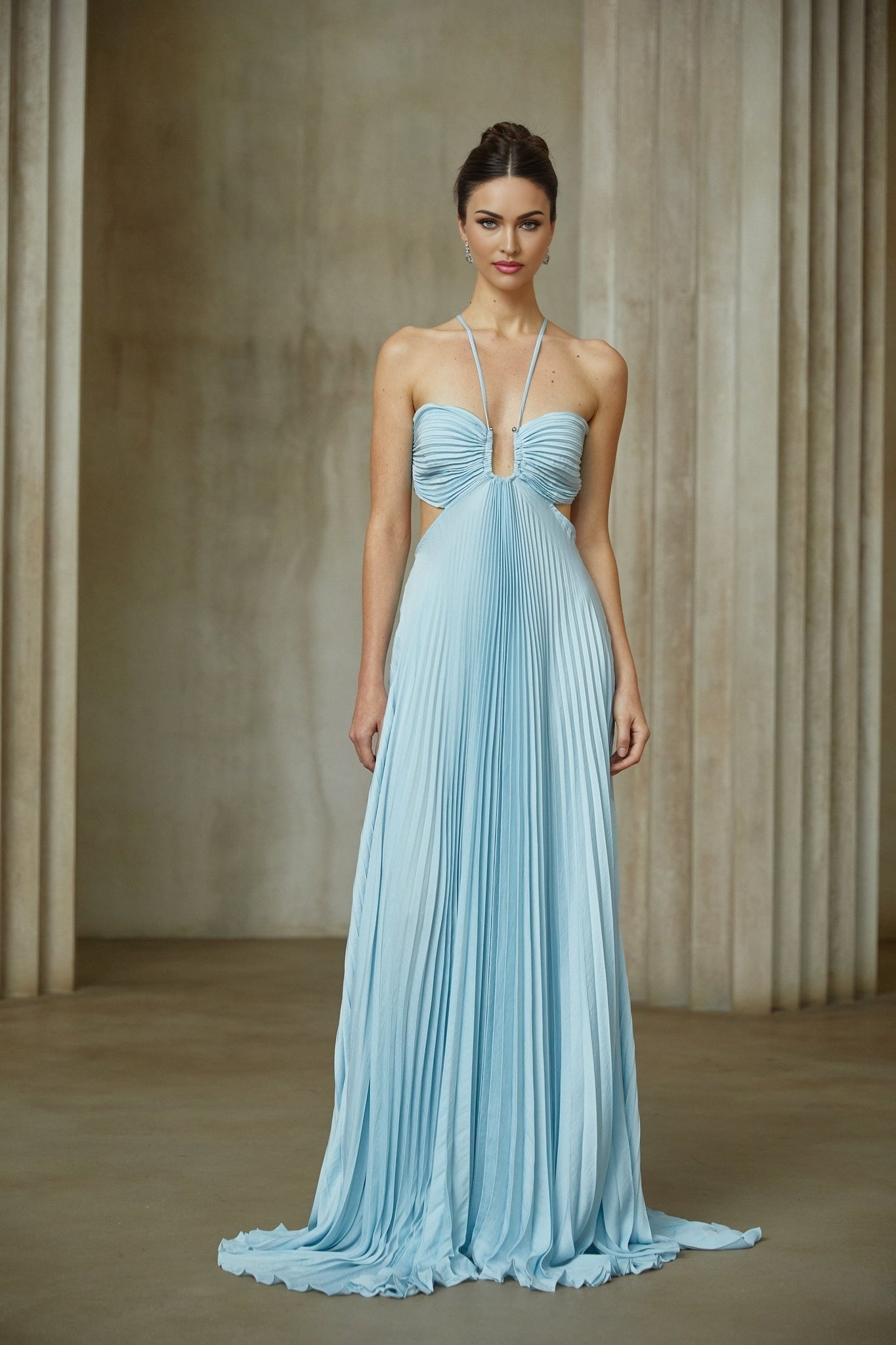 A woman wearing a long blue pleated maxi dress with a cutout detail standing against a plain background