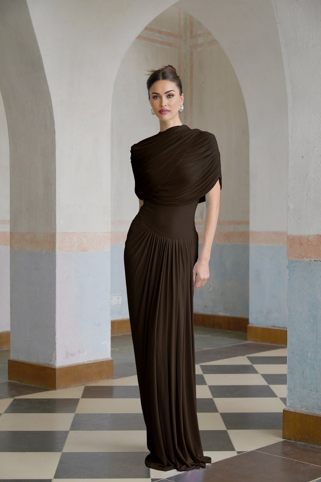 A woman wearing a long brown asymmetric dress with pleated details standing in a relaxed pose
