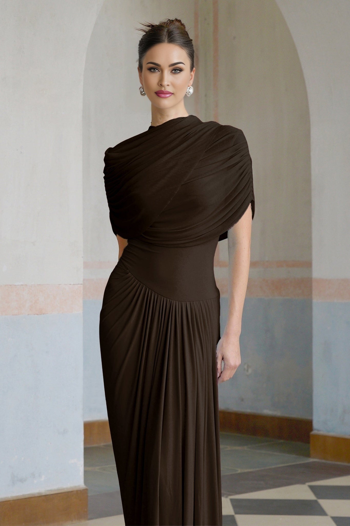 A woman wearing a long brown asymmetric pleated maxi dress with one shoulder strap the dress flows elegantly to the floor
