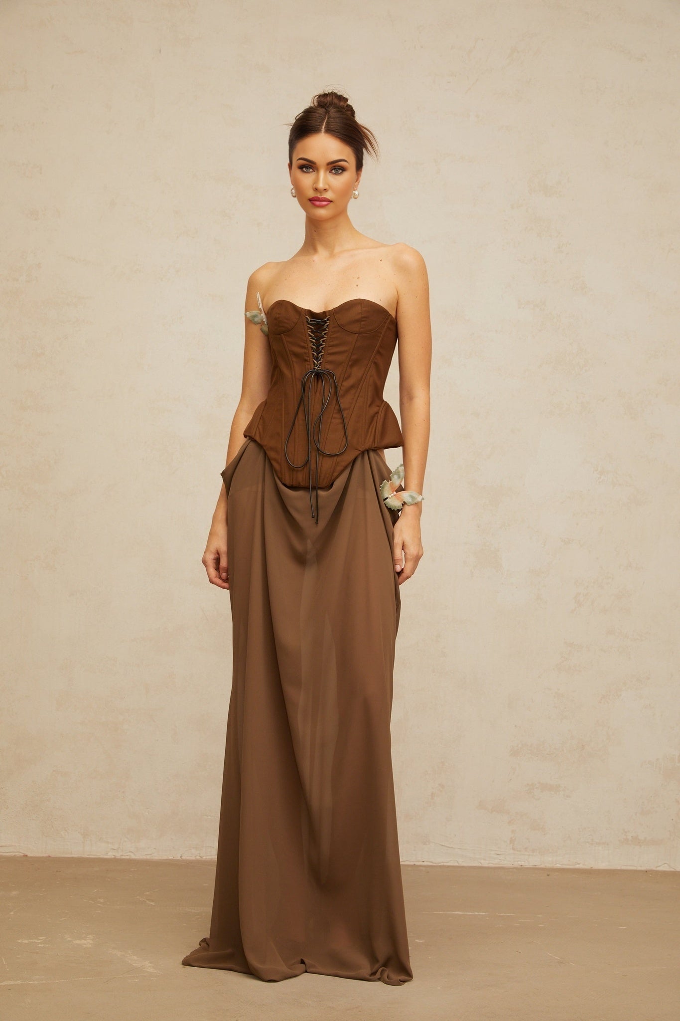 A woman wearing a long brown maxi dress with a corset style top and butterfly pin detail at the waist