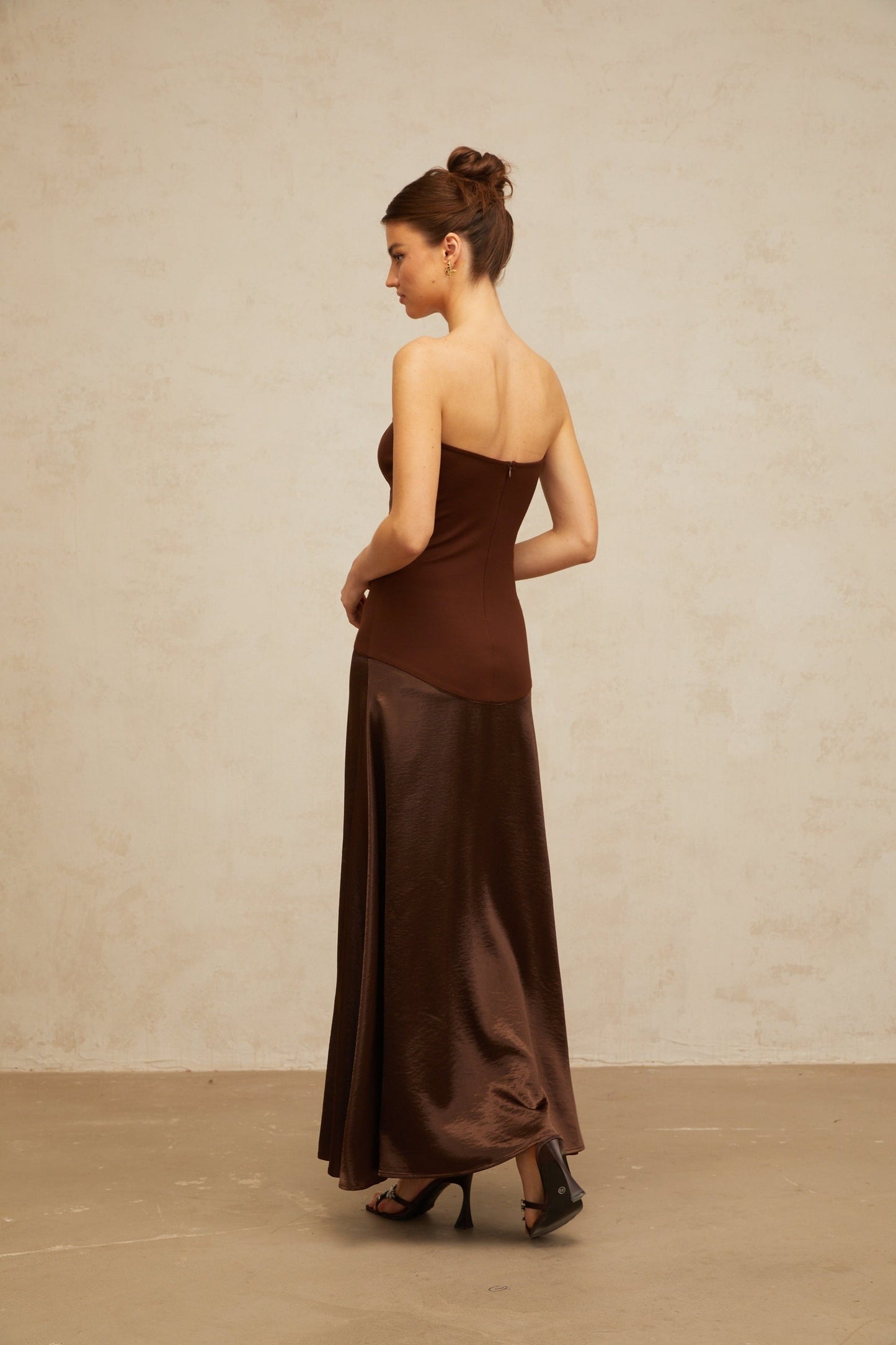 A woman wearing a long brown offshoulder dress with satin panels standing in a relaxed pose