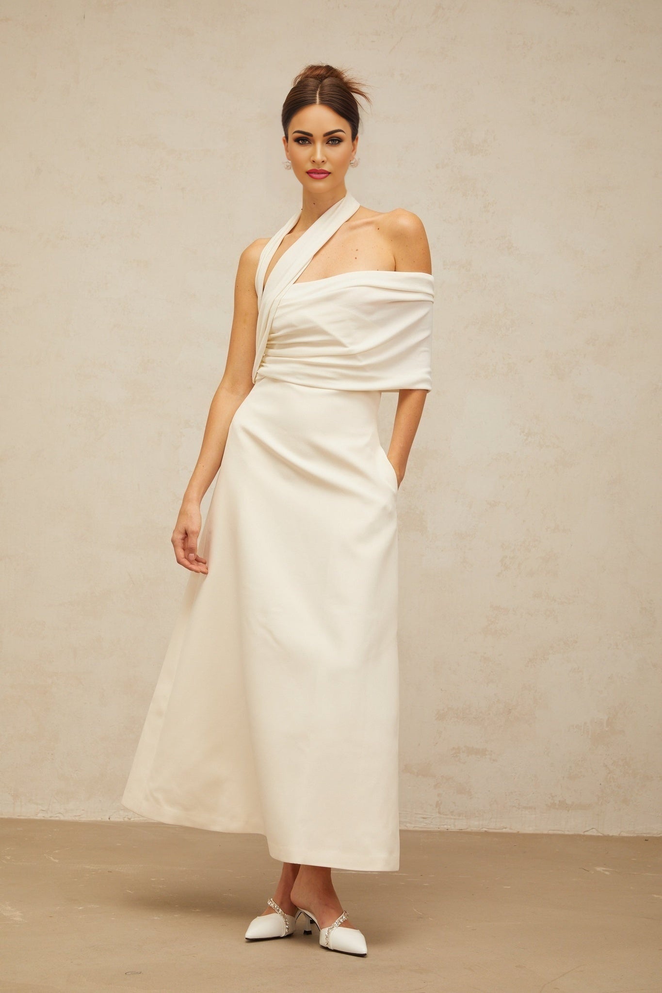 A woman wearing a long creamwhite maxi dress with an asymmetric design standing in a neutral background
