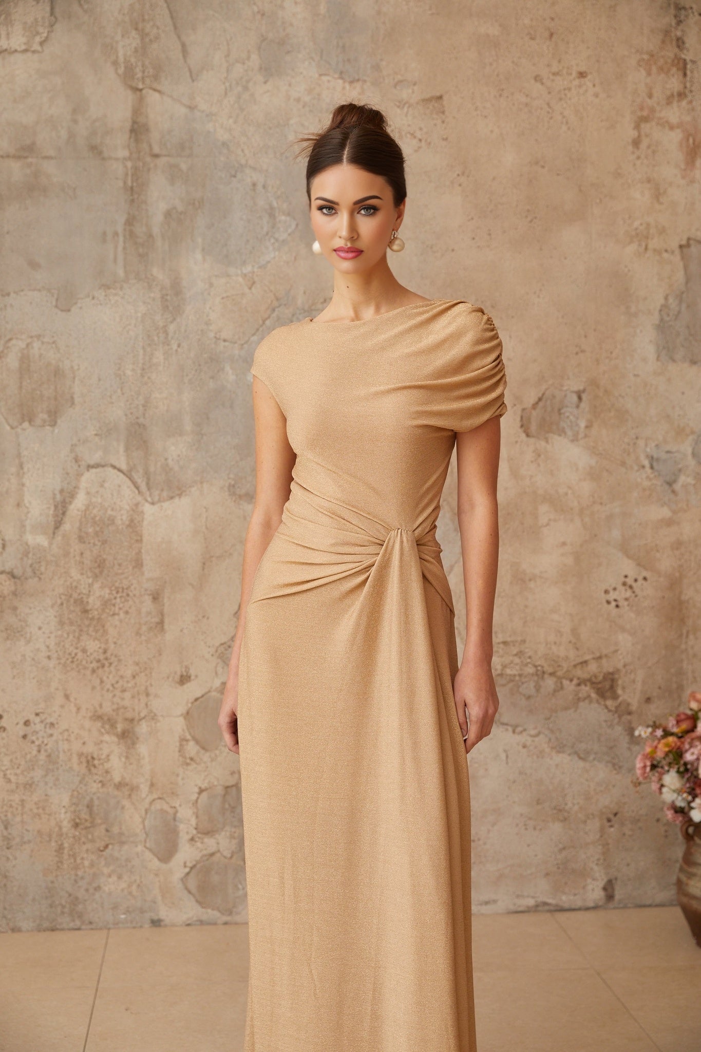 A woman wearing a long flowing gold maxi dress with an asymmetrical neckline and pleated skirt standing in an elegant pose