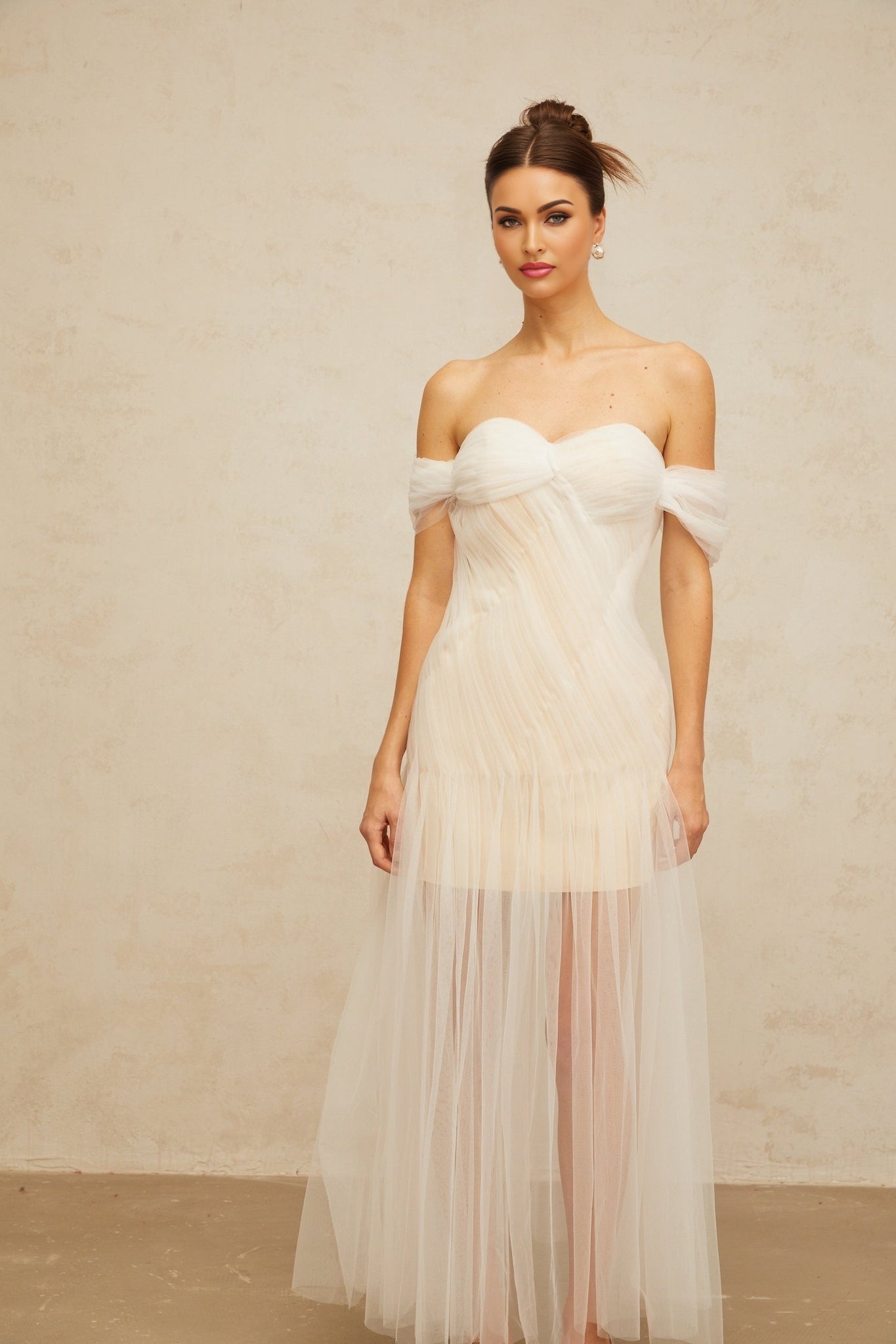 A woman wearing a long flowing off white maxi dress with pleated tulle fabric and delicate details standing in an elegant pose
