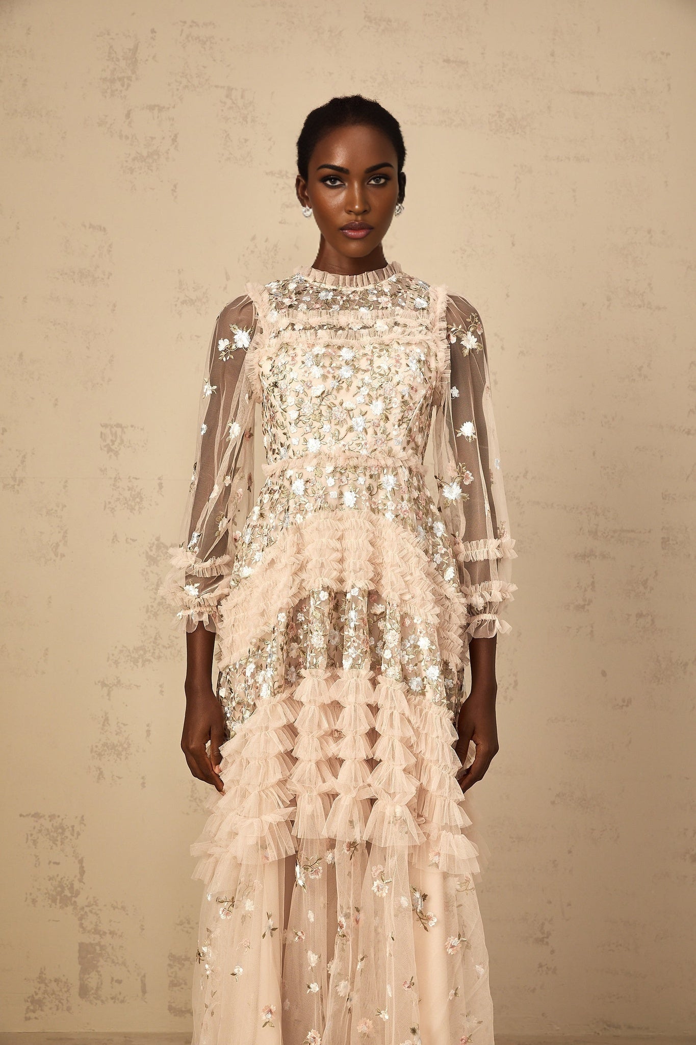 A woman wearing a long flowy maxi dress with floral embroidery and tiered tulle skirt in a soft pastel color standing against a plain background