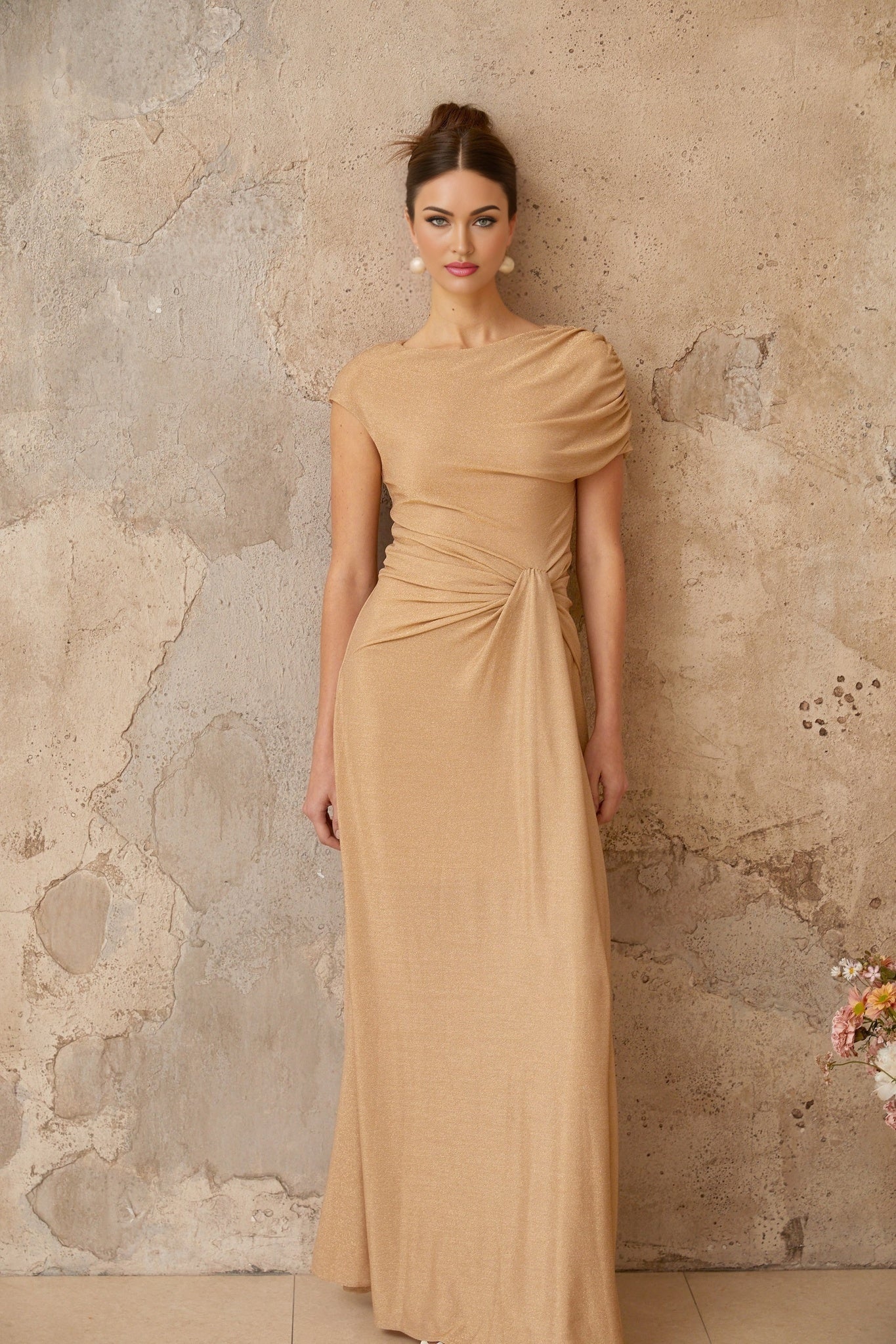 A woman wearing a long gold maxi dress with an asymmetrical neckline and pleated skirt standing in an elegant pose