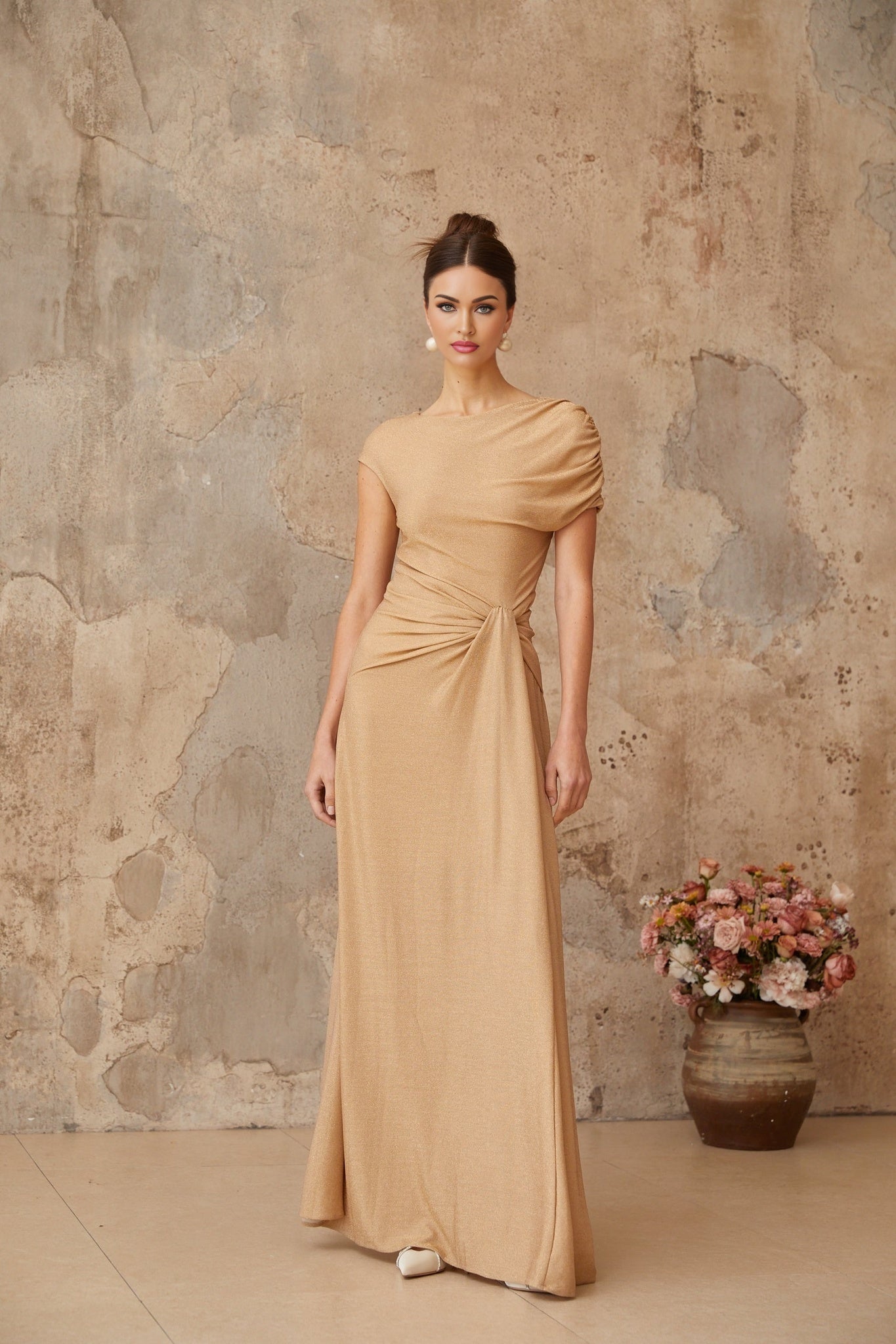 A woman wearing a long gold maxi dress with asymmetrical pleating and a flowing skirt standing in an elegant pose