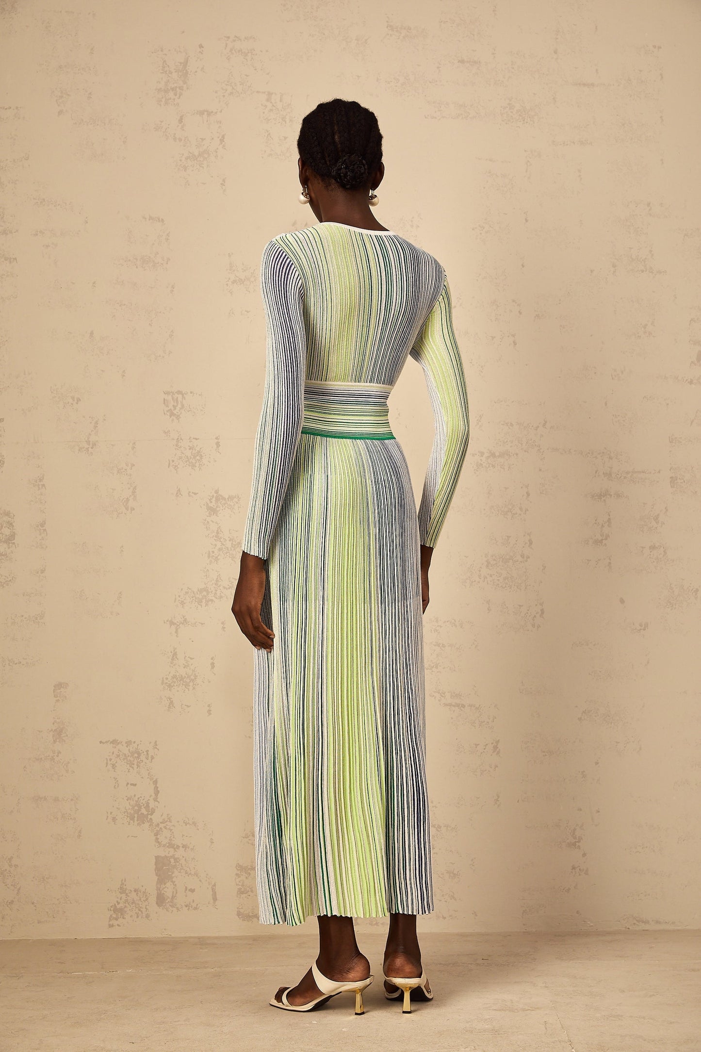 A woman wearing a long green maxi dress with long sleeves and sequin stripes standing in a neutral background