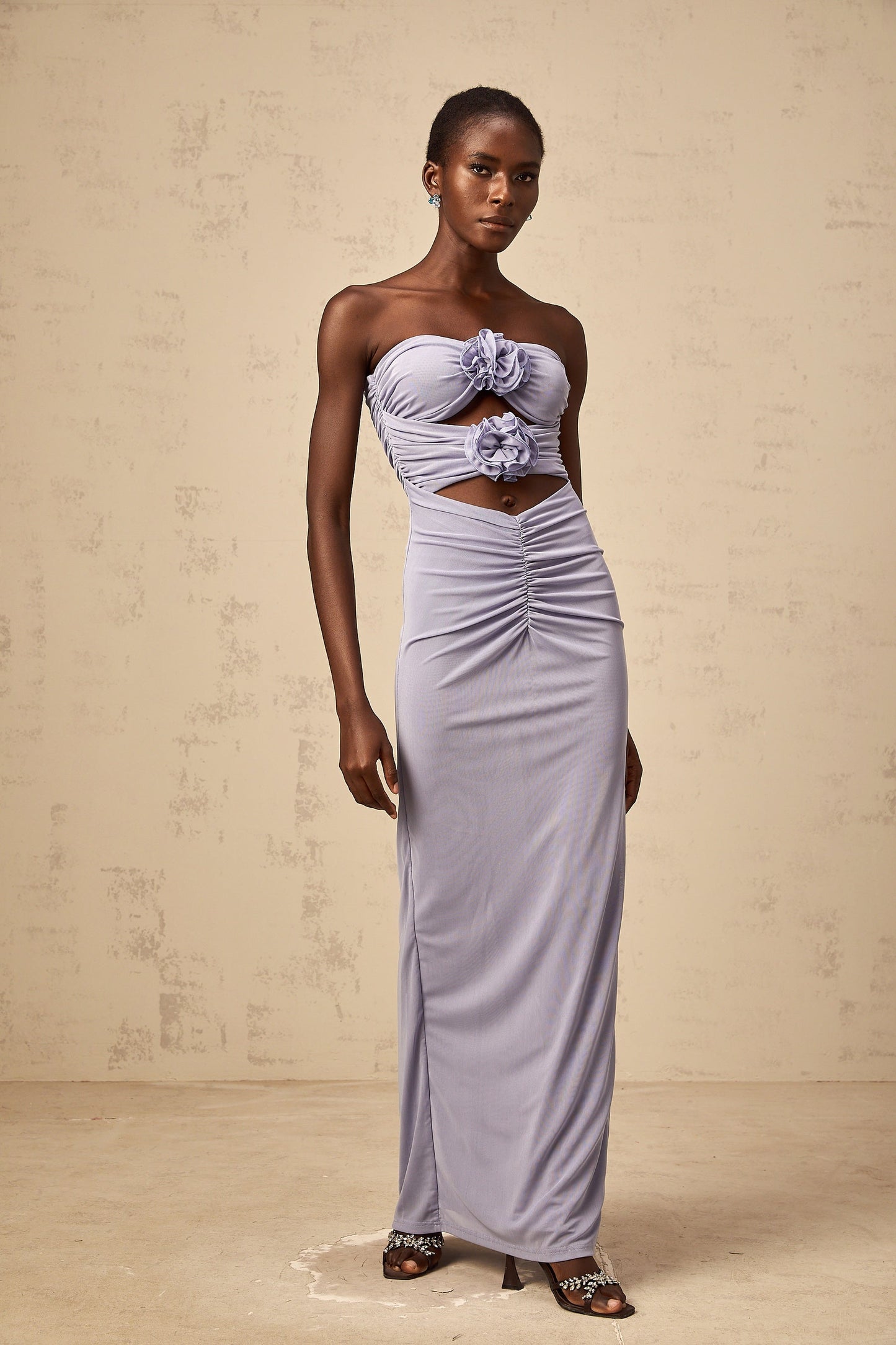 A woman wearing a long lavender dress with a fitted corset bodice and ruched detailing featuring 3D petal embellishments along the neckline and skirt