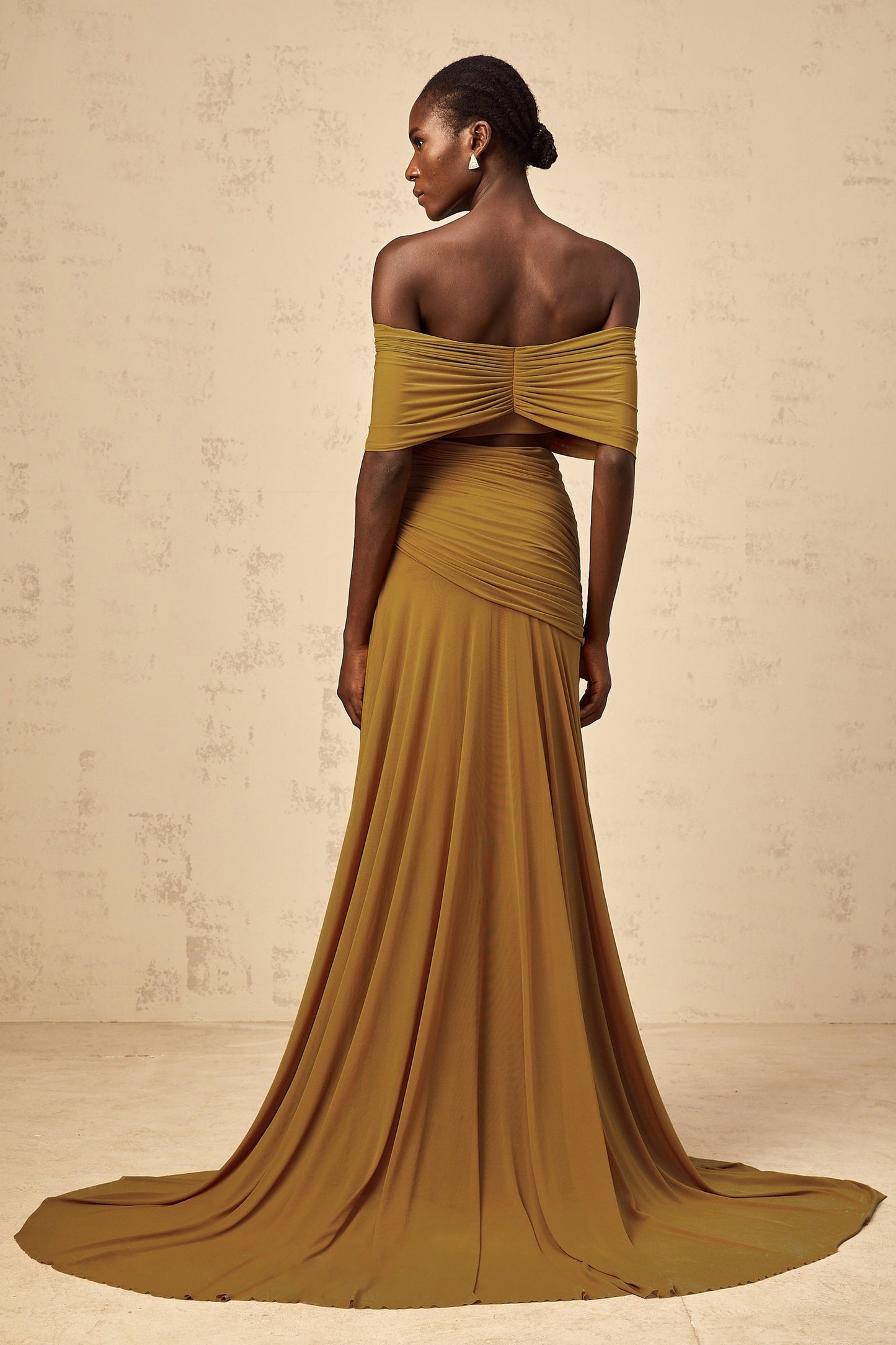 A woman wearing a long ocher colored dress with offshoulder design ruched details and cut out sections The dress has a maxi length and flows elegantly