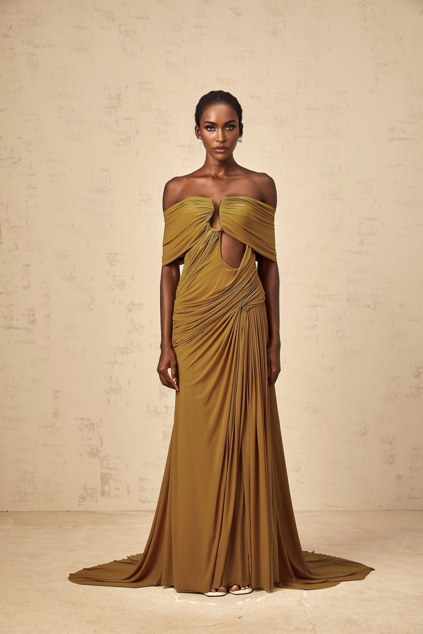 A woman wearing a long ocher colored maxi dress with offshoulder design and cutout details The dress features ruching and a flowy silhouette
