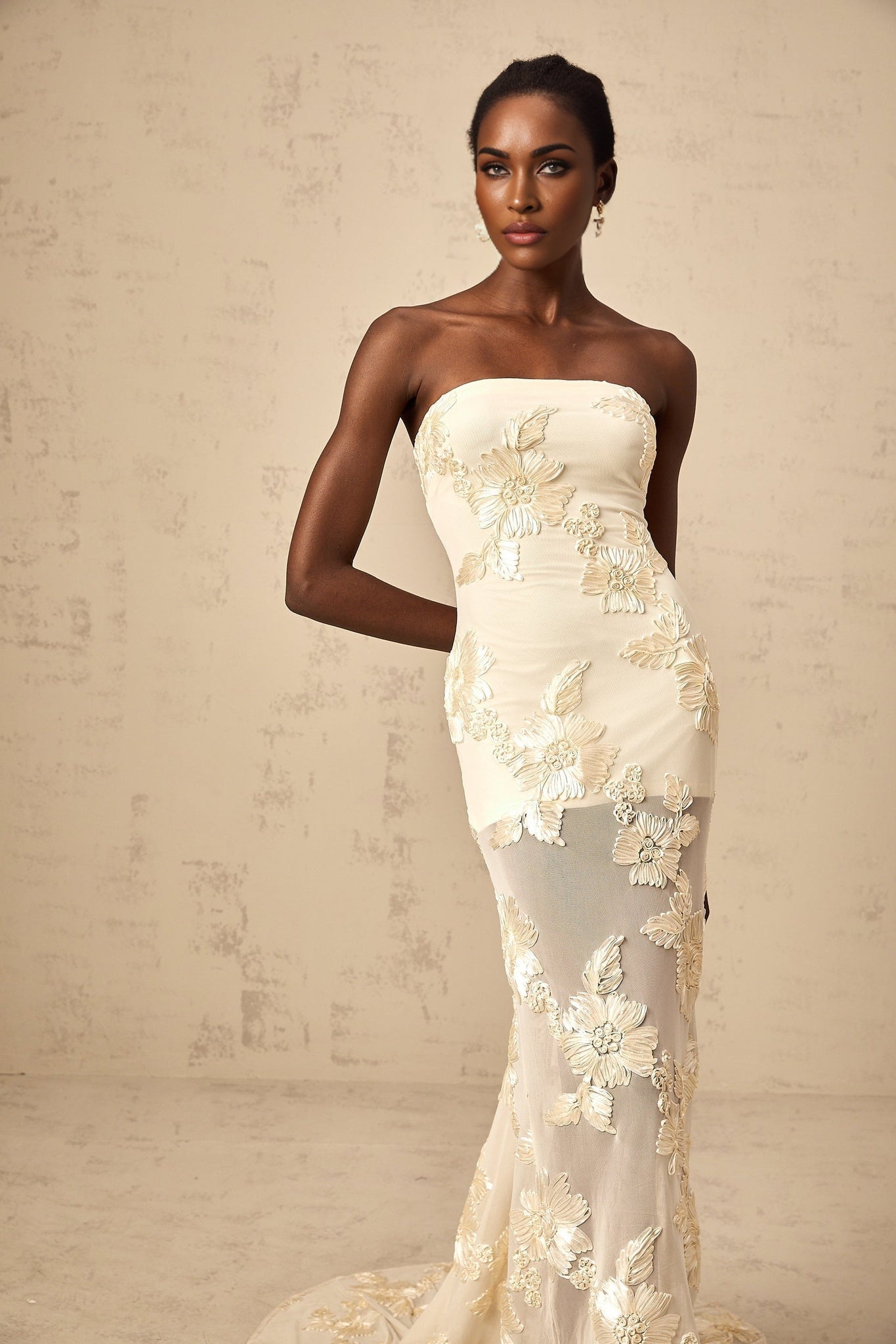 A woman wearing a long offshoulder maxi dress in neutral colors with embroidered details standing against a plain background