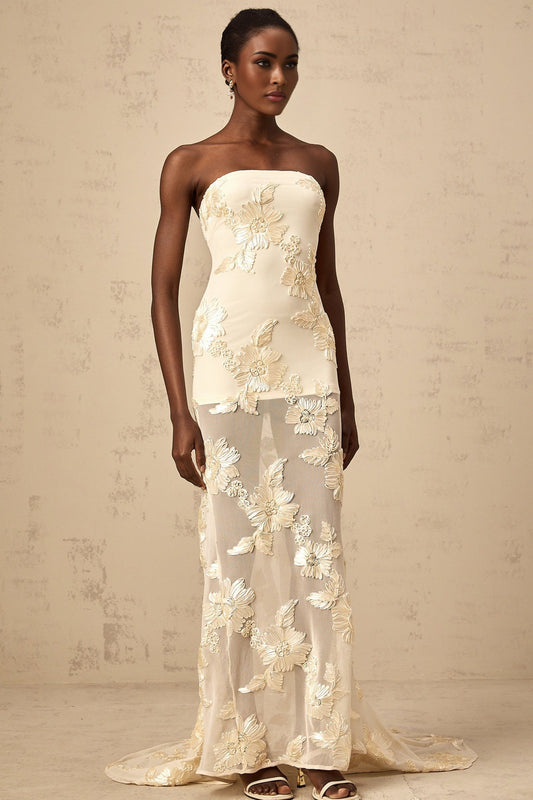 A woman wearing a long offshoulder maxi dress in neutral tones with embroidered details standing against a plain background