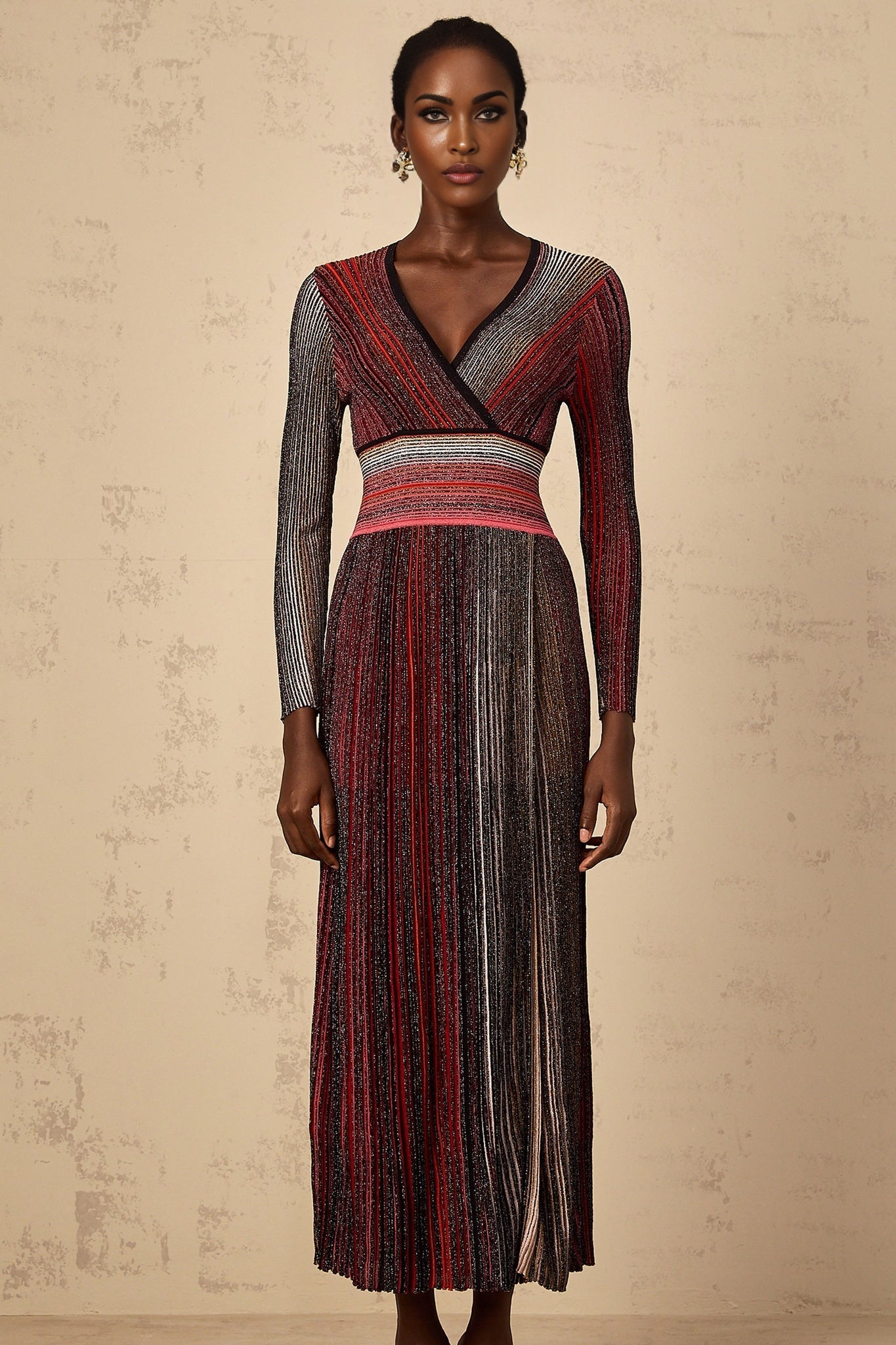 A woman wearing a long red maxi dress with long sleeves and vertical sequin stripes standing against a plain background