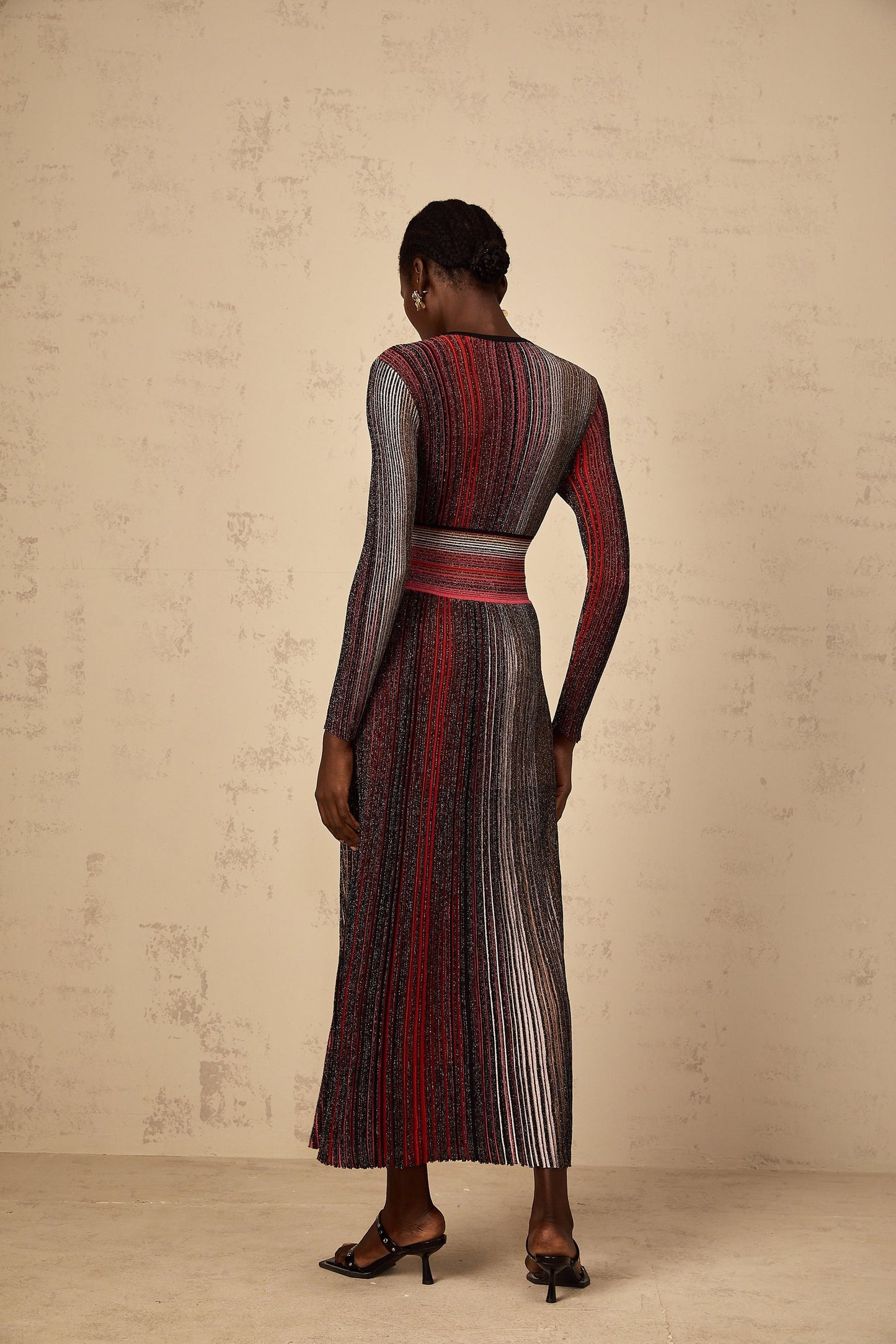 A woman wearing a long red maxi dress with long sleeves and vertical sequin stripes standing against a plain background