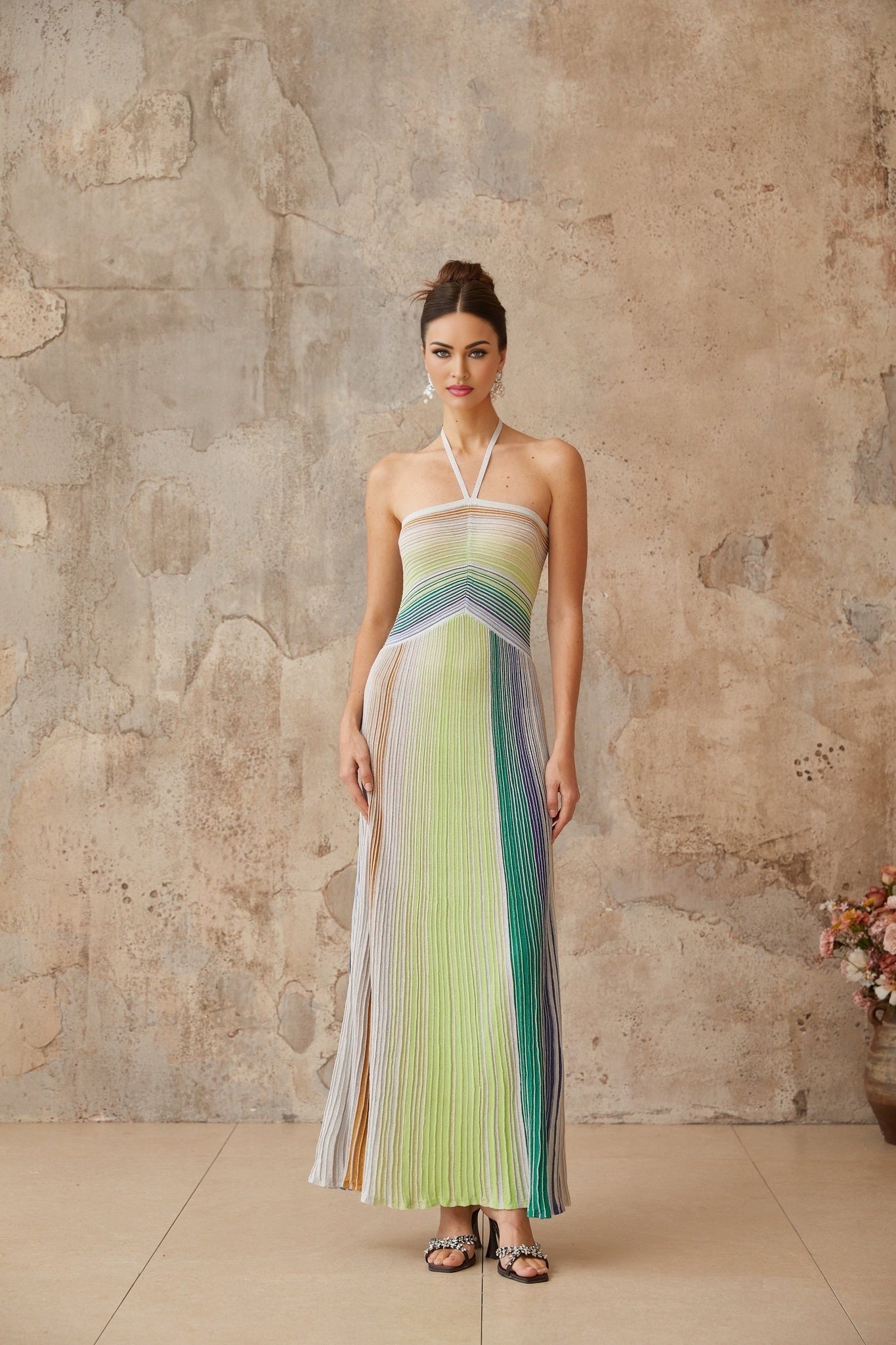 A woman wearing a long shimmering metallic knit maxi dress with a backless design standing in an elegant pose