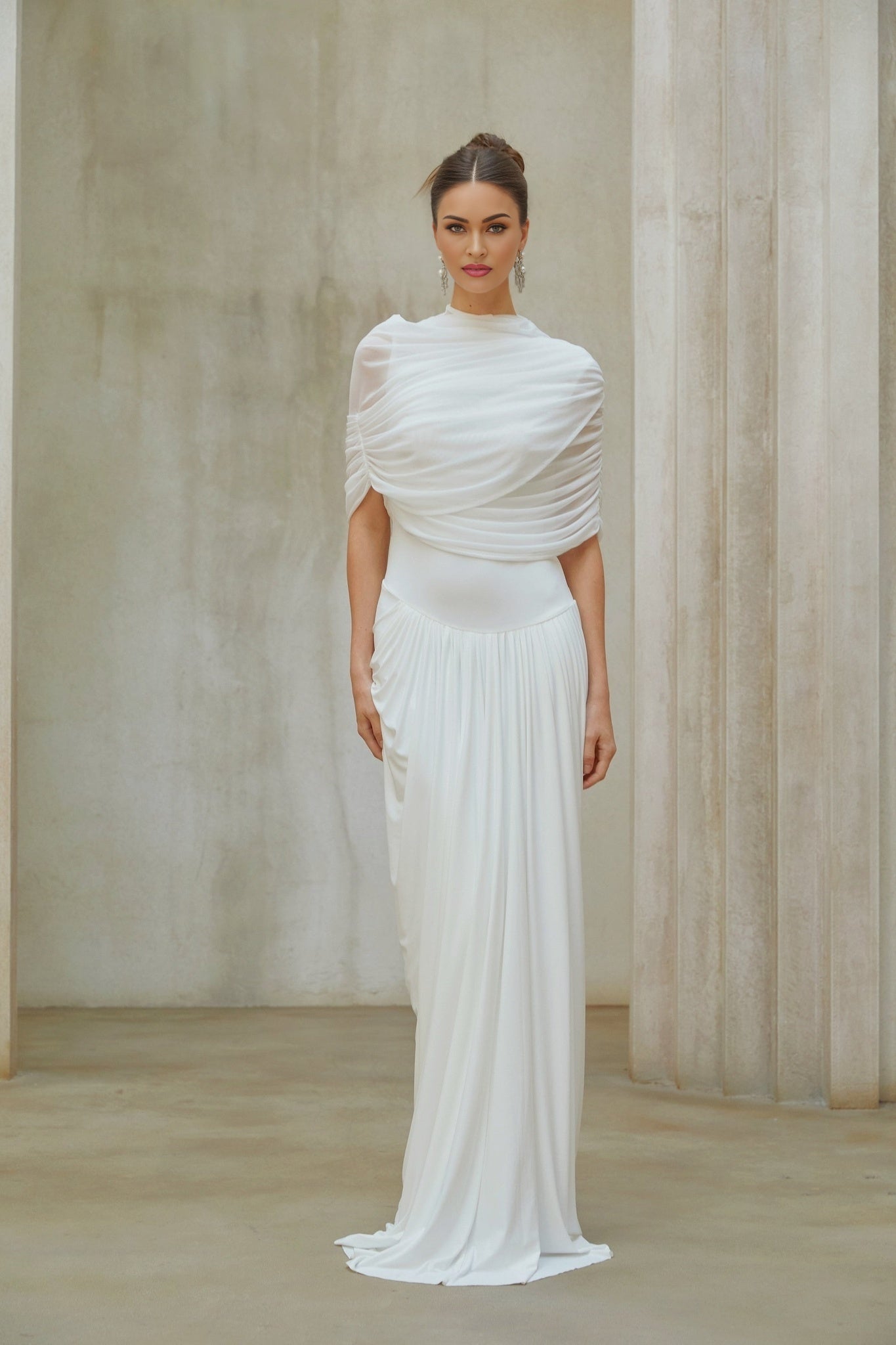 A woman wearing a long white asymmetrical maxi dress with pleated details standing in a neutral background