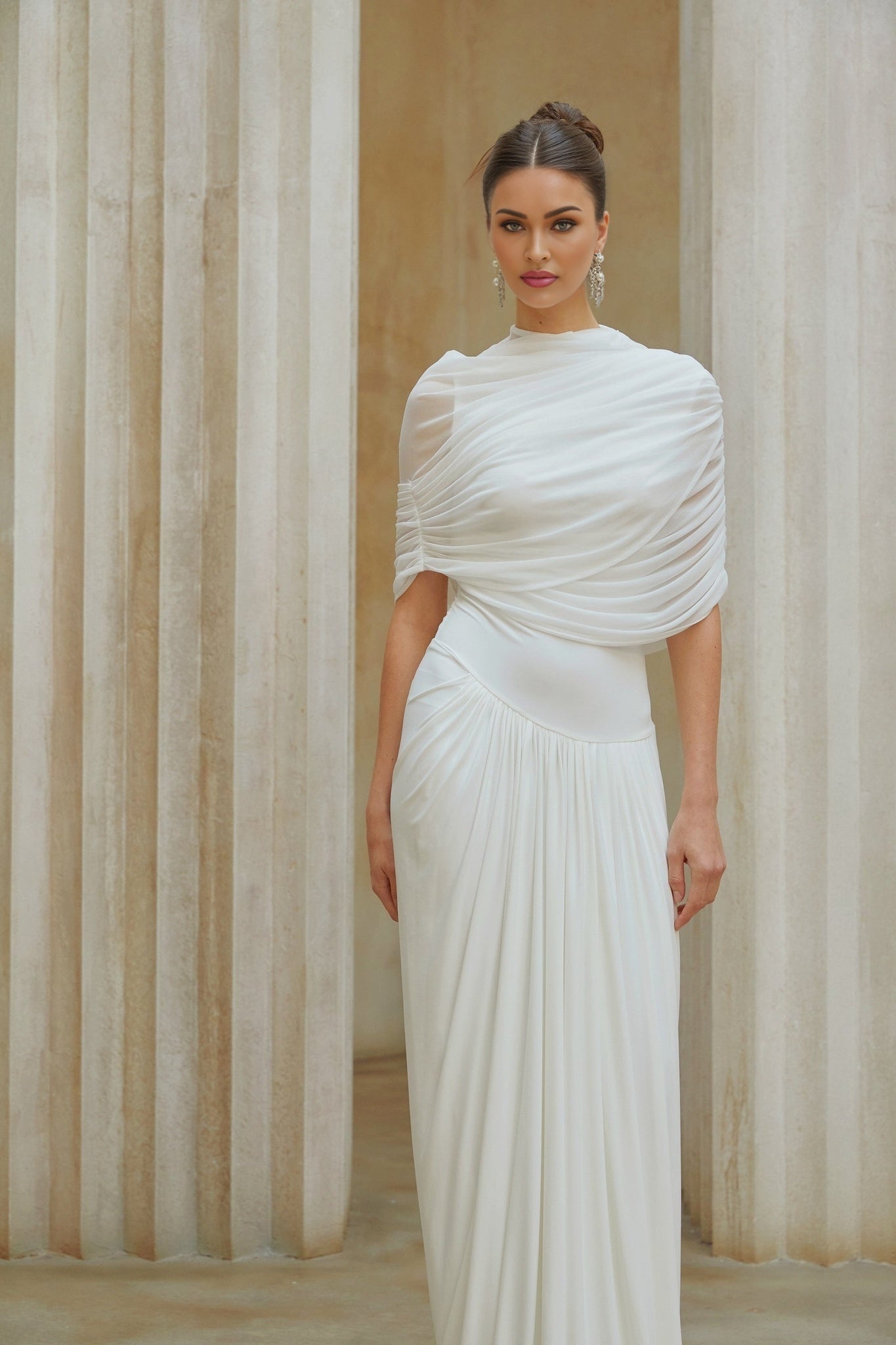 A woman wearing a long white asymmetrical maxi dress with pleated details standing in a neutral setting