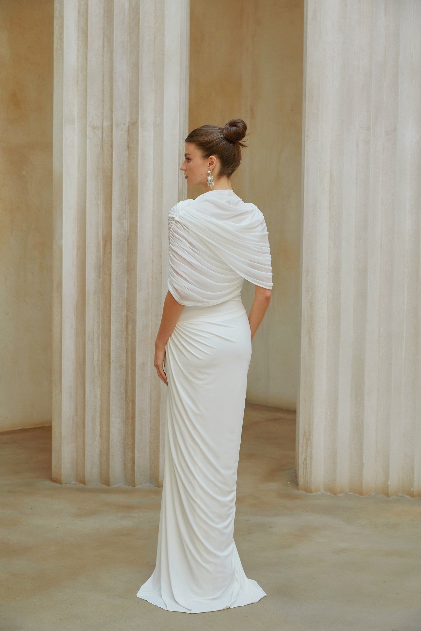 A woman wearing a long white asymmetrical maxi dress with pleated details standing in a relaxed pose