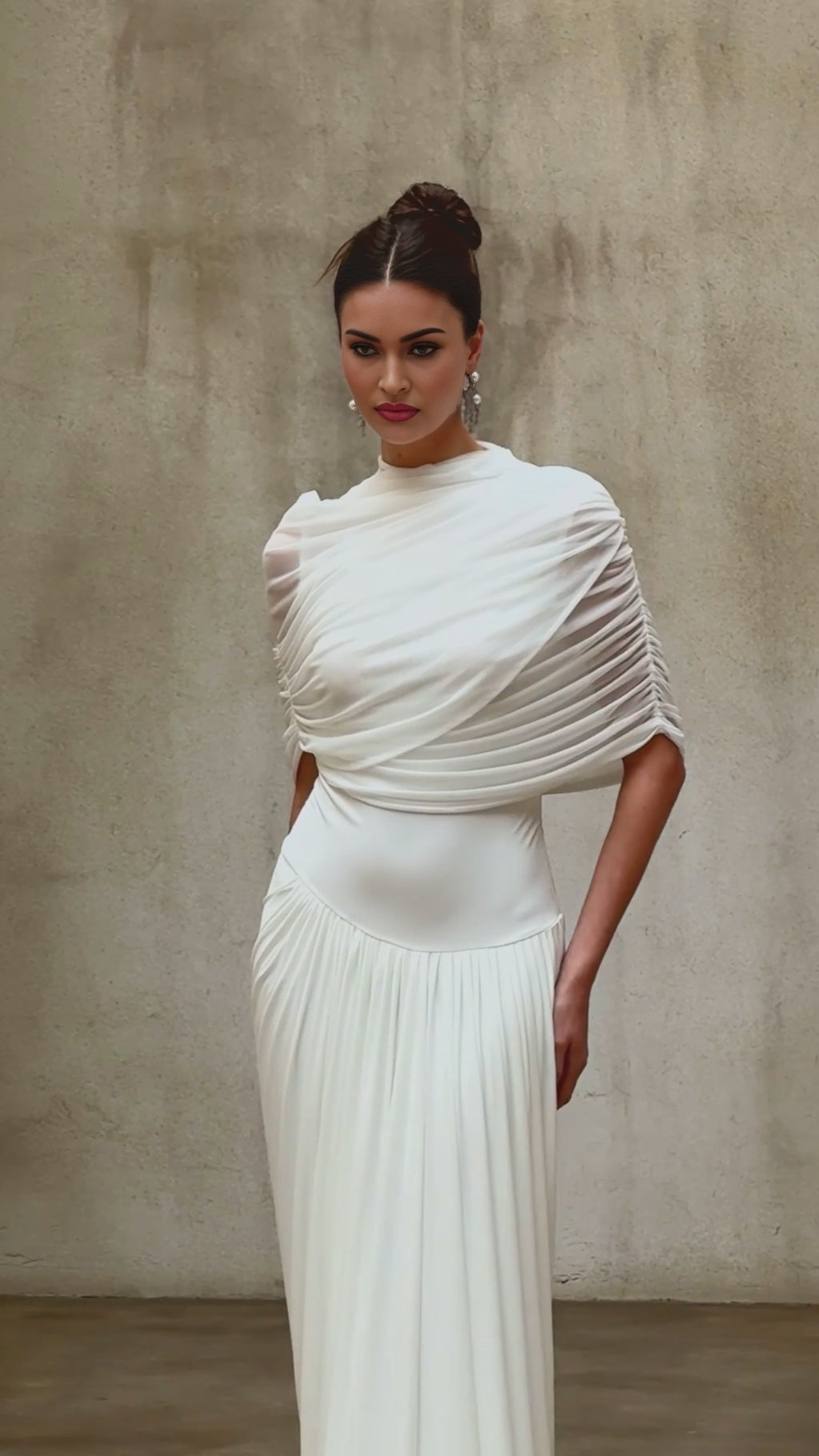 A woman wearing a long white asymmetrical maxi dress with pleated details standing in a relaxed pose