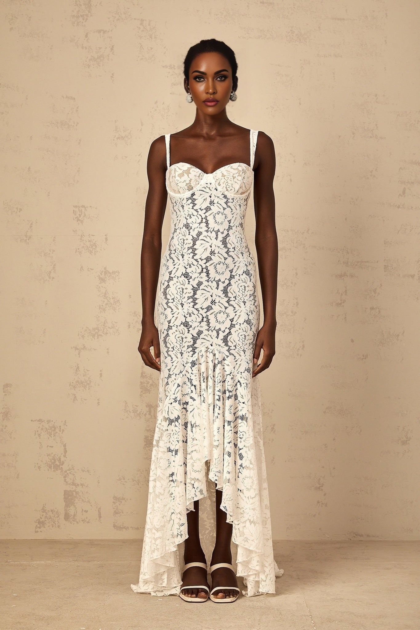 A woman wearing a long white maxi dress with floral lace detailing and a bustier style top standing against a plain background