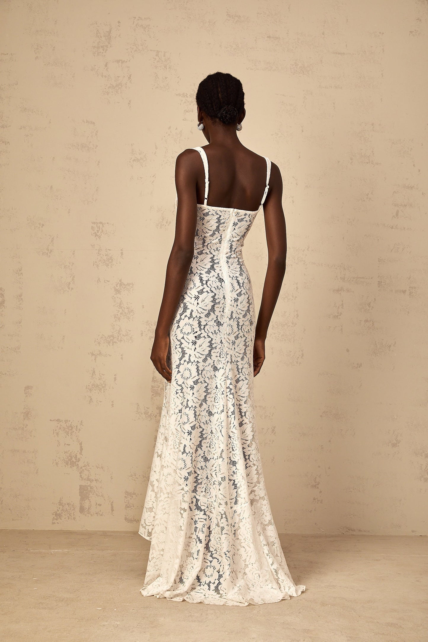A woman wearing a long white maxi dress with floral lace details and a bustier top standing against a plain background