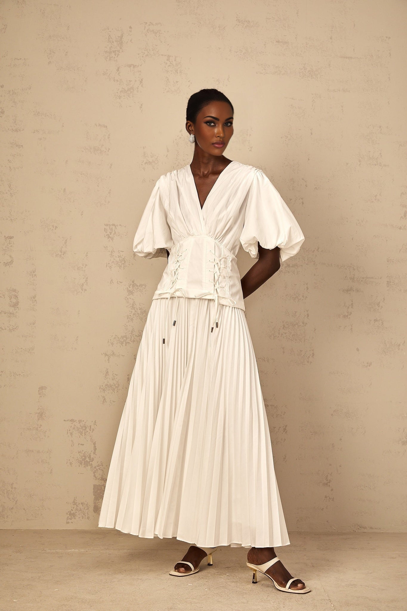 A woman wearing a long white pleated maxi dress with lace up details standing in a relaxed pose