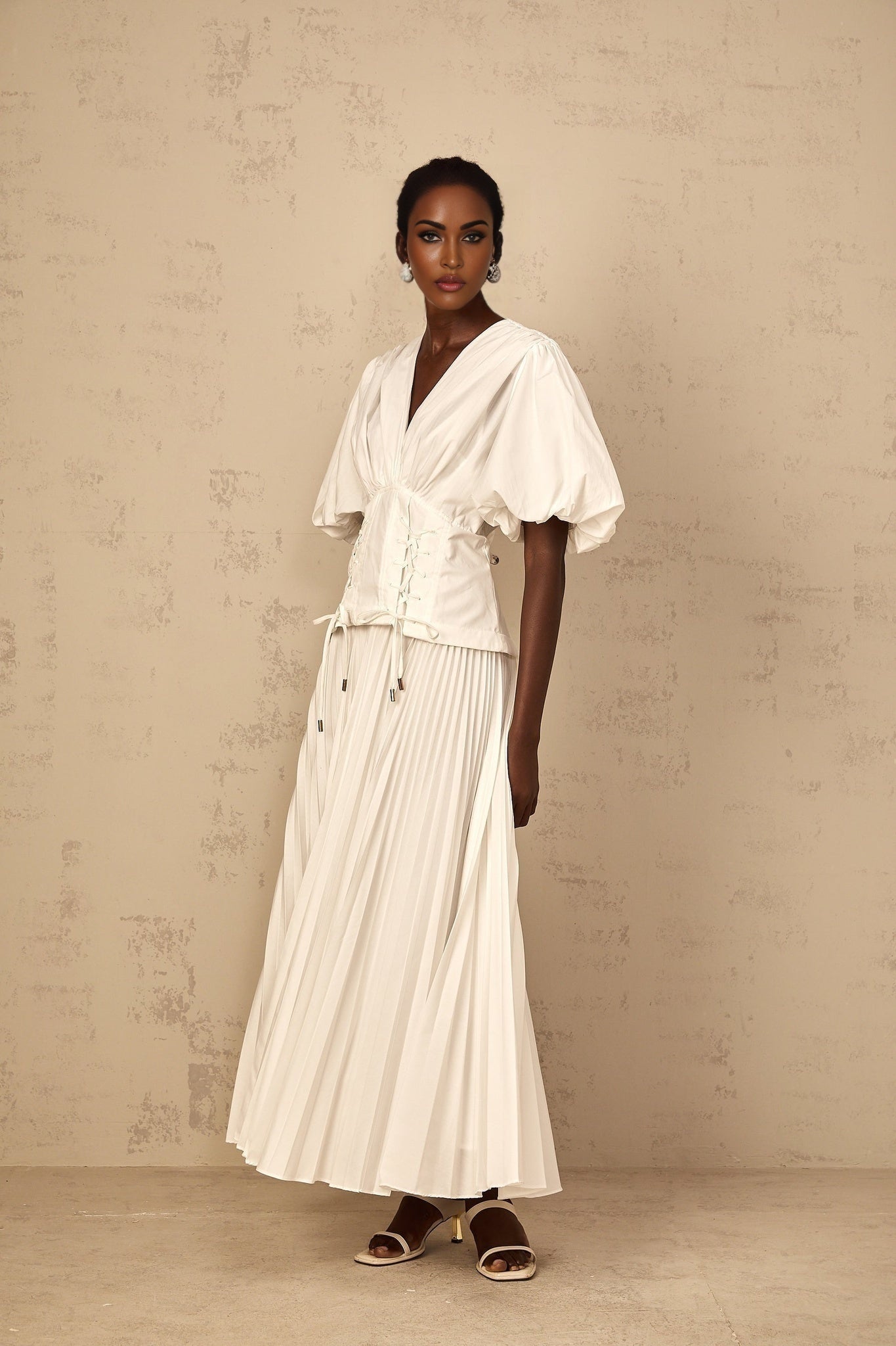 A woman wearing a long white pleated maxi dress with lace up details standing in a relaxed pose
