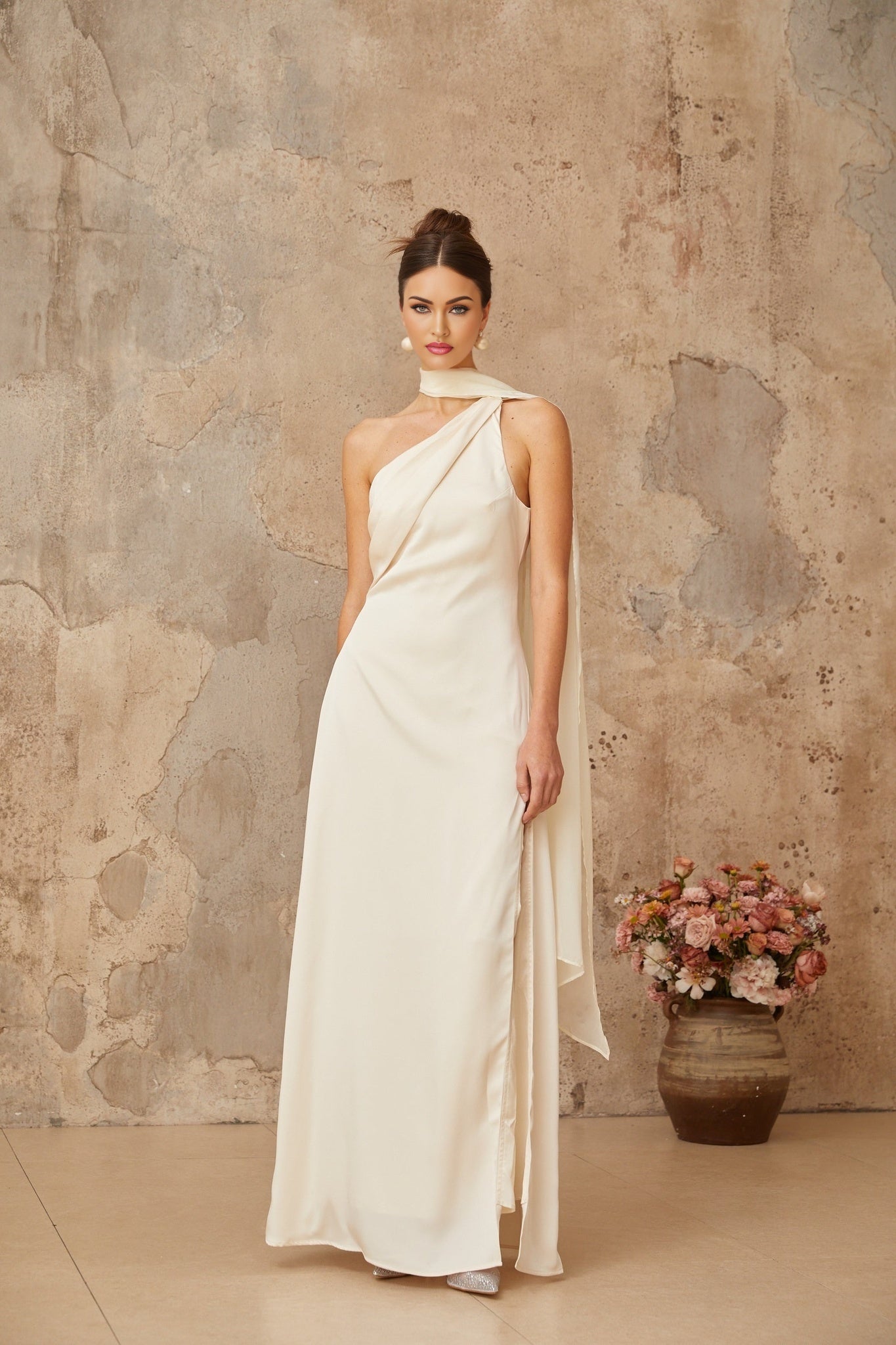 A woman wearing a long white satin maxi dress with a scarflike design at the neckline standing against a plain background