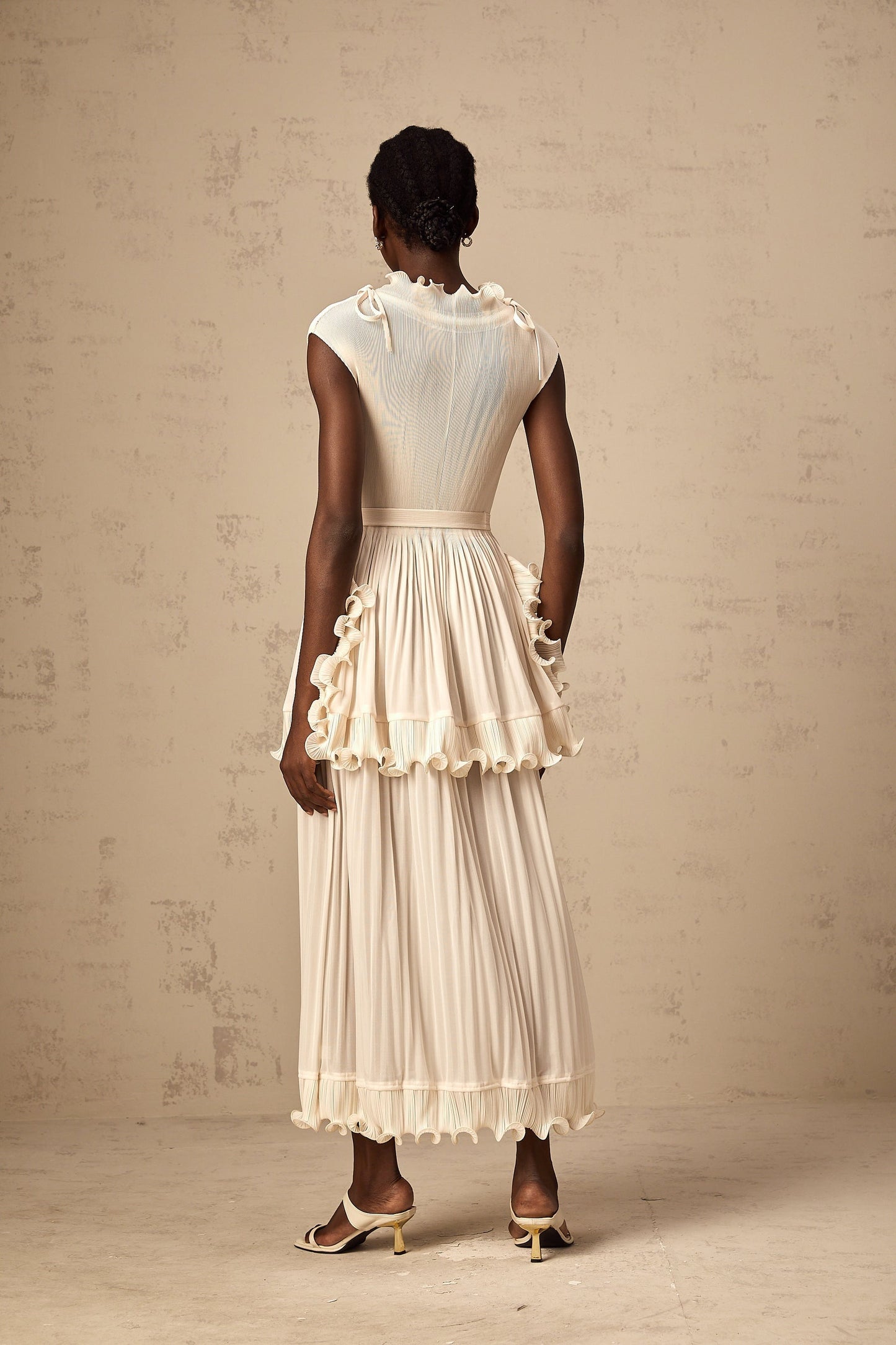 A woman wearing a long white sleeveless maxi dress with ruffle details and pleats standing against a plain background