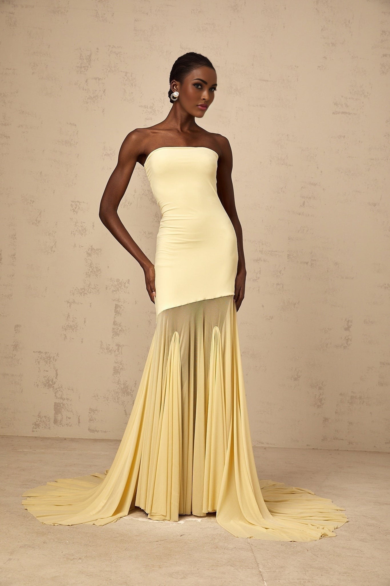 A woman wearing a long yellow offshoulder maxi dress made of sheer tulle fabric posing against a plain background