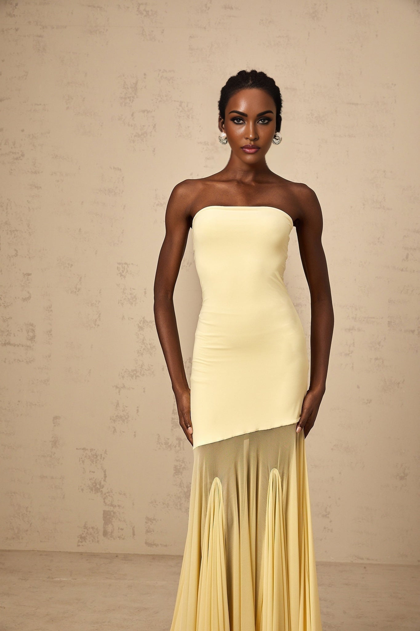 A woman wearing a long yellow offshoulder maxi dress with sheer tulle fabric standing against a plain background