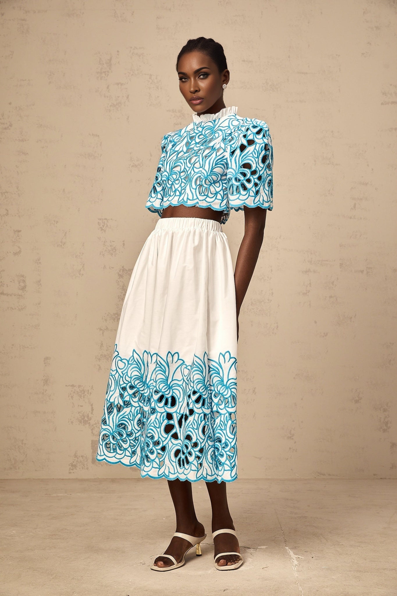 A woman wearing a matching blue twopiece outfit consisting of a sleeveless crop top with cutout details and embroidery paired with a high waisted skirt featuring similar embroidery and cutout designs