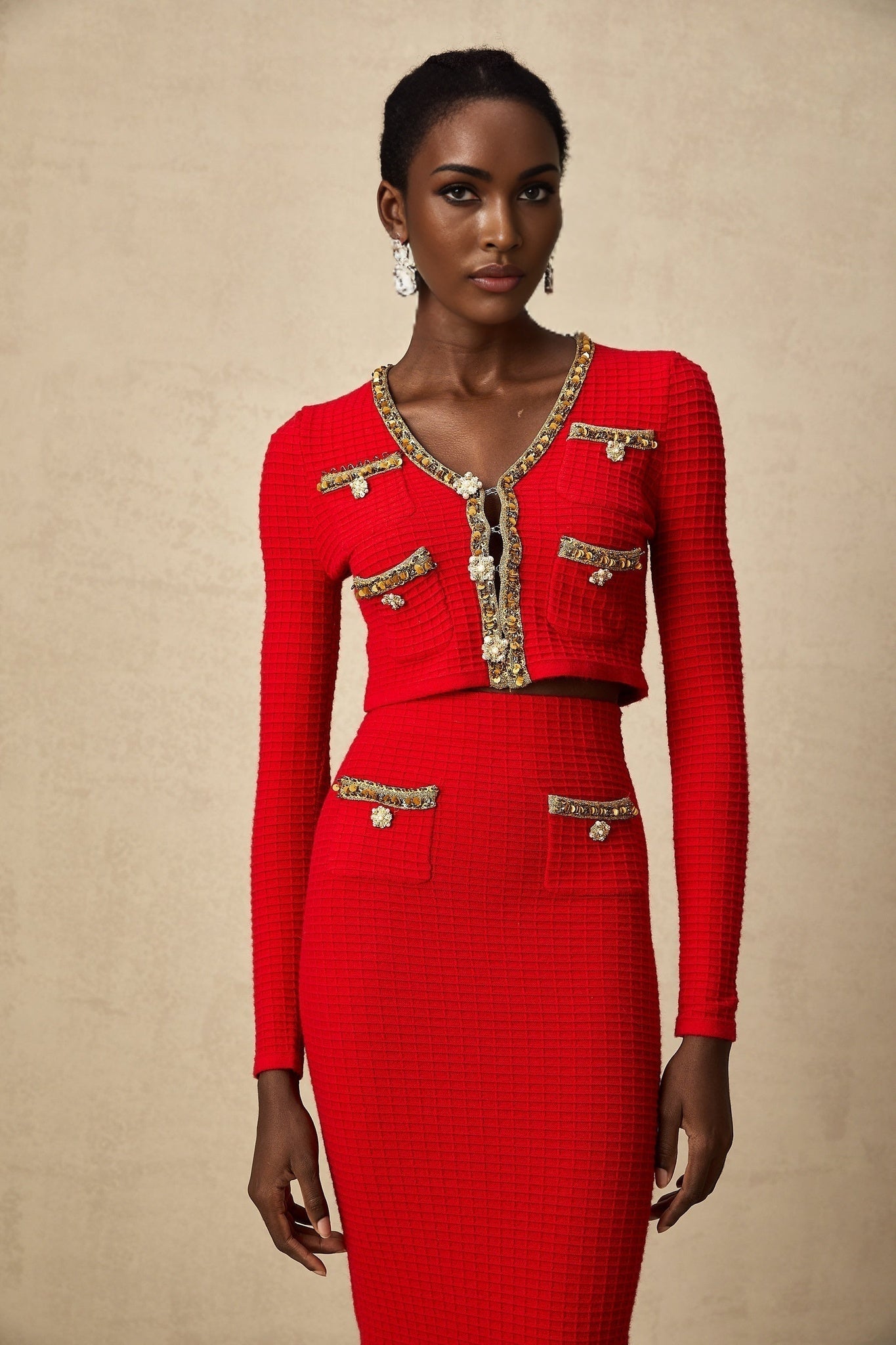 A woman wearing a matching red knitted cardigan and skirt set with embellishments The cardigan has long sleeves and the skirt is knee length Both pieces feature intricate embroidery or beadwork details