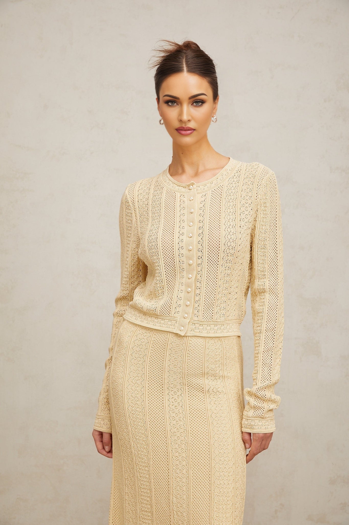 A woman wearing a matching white pointelle knit cardigan and skirt set The cardigan has long sleeves and an open front The skirt is knee length The set has a delicate lacy pattern throughout