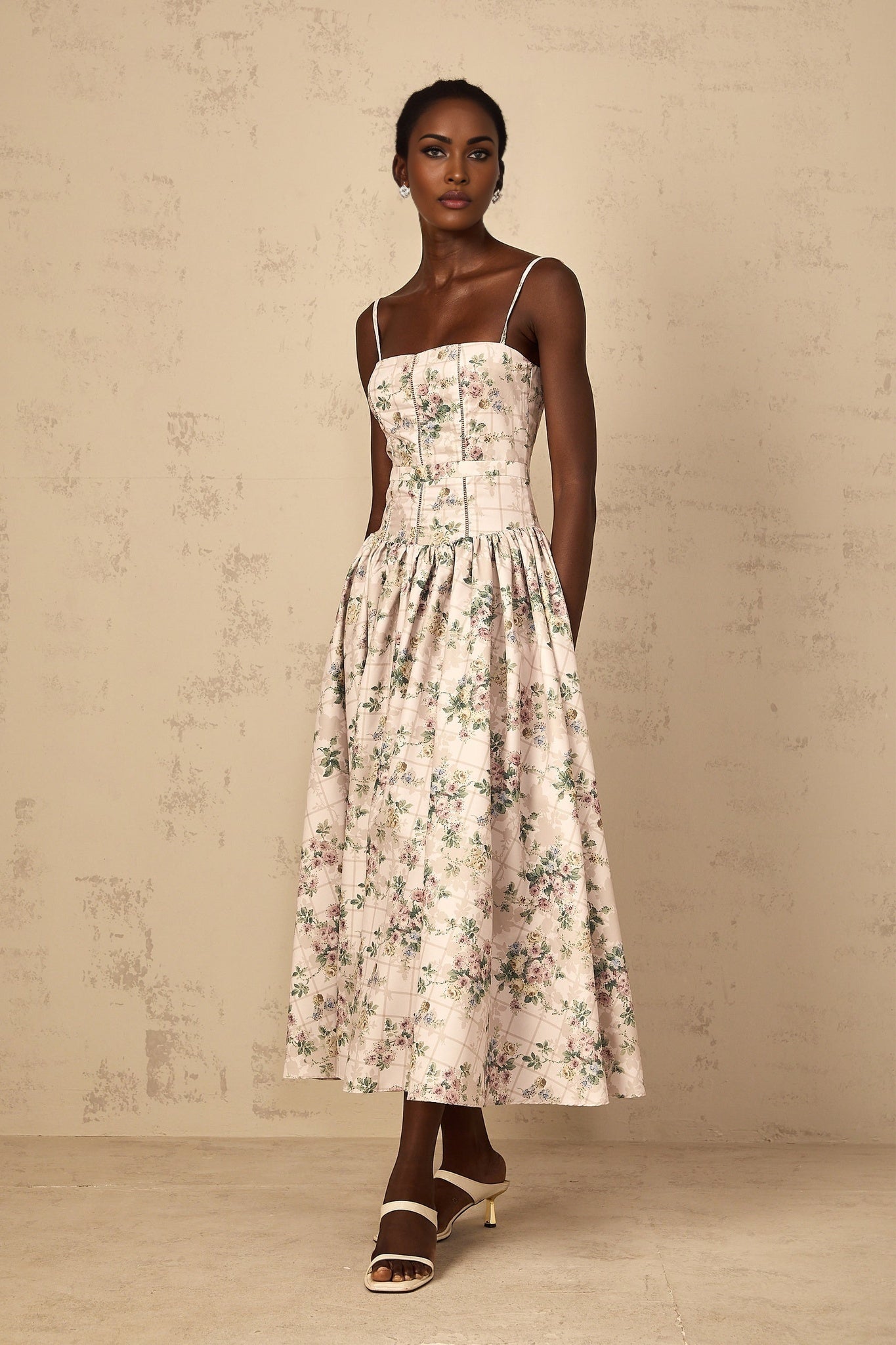 A woman wearing a midi dress with a neutraltoned floral print standing against a plain background