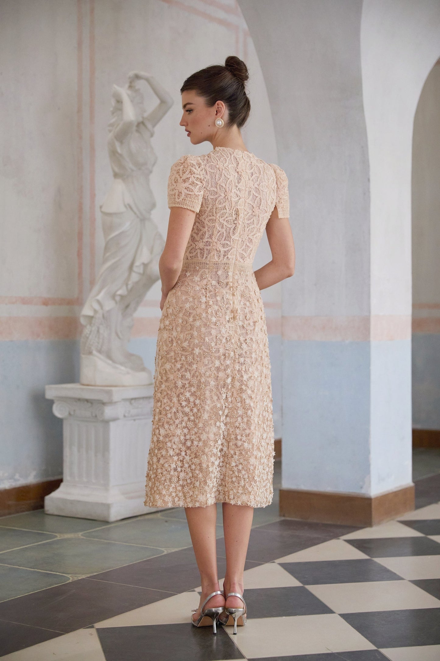 A woman wearing a neutral colored midi dress with a crochet pattern and faux flower details standing in a relaxed pose