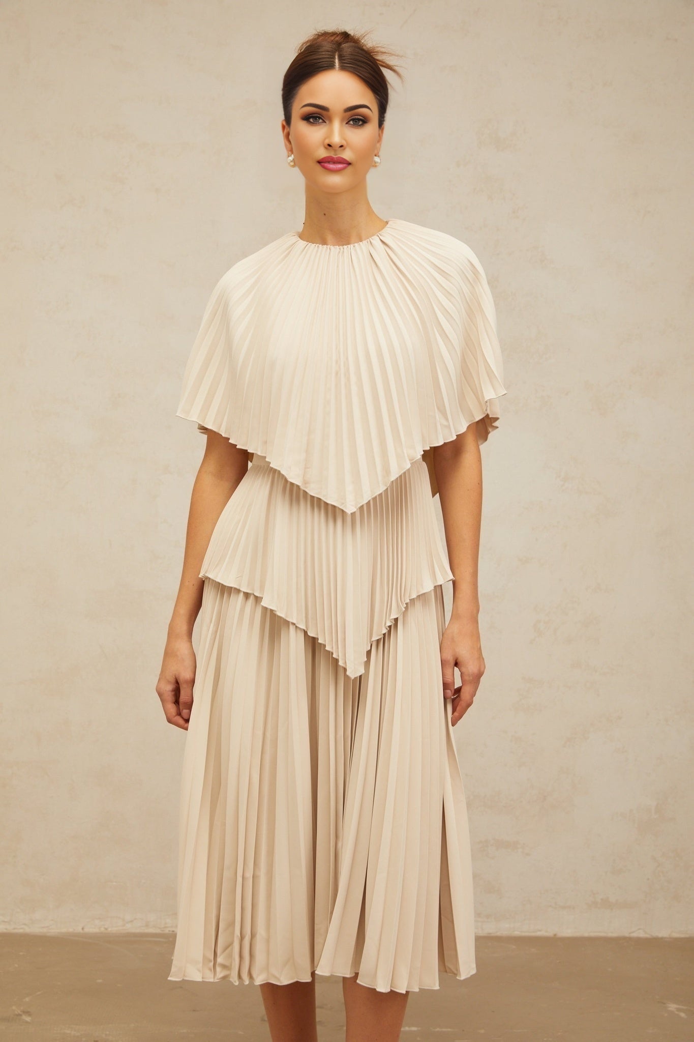 A woman wearing a neutralcolored cape style midi dress with pleated details standing in a relaxed pose