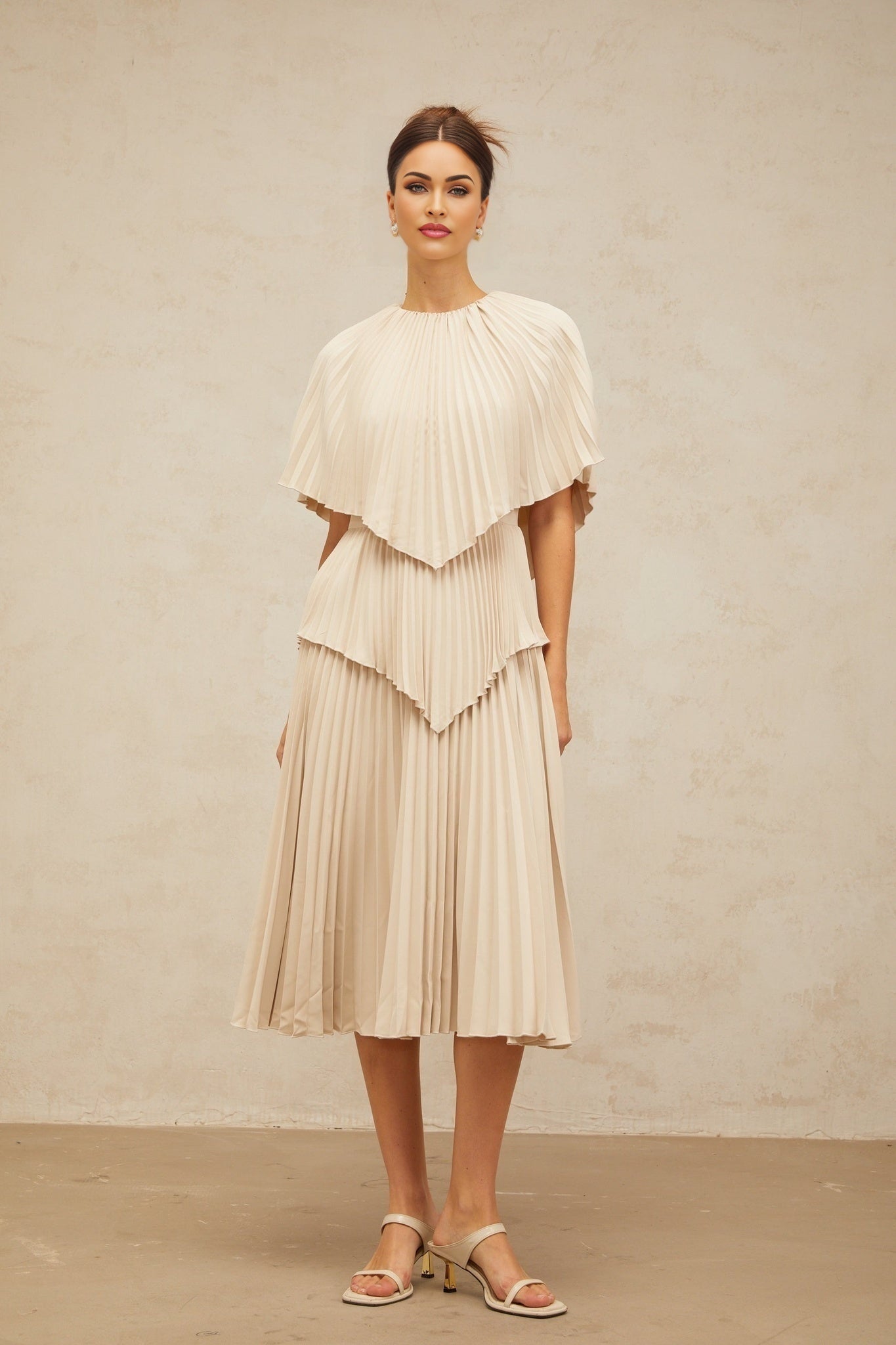 A woman wearing a neutralcolored cape style pleated midi dress with flowing fabric and a sophisticated silhouette