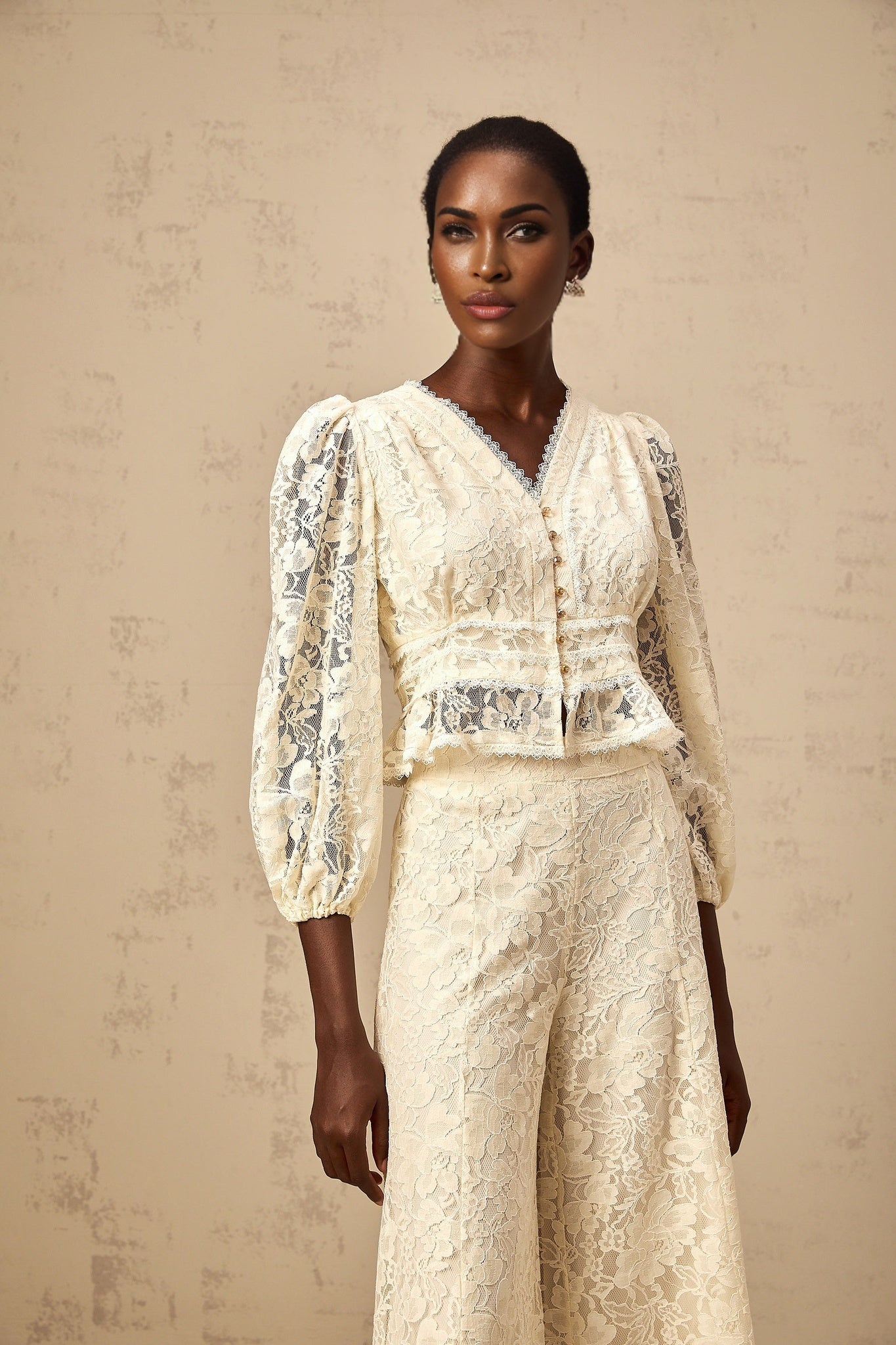 A woman wearing a neutralcolored matching set consisting of a floral lace blouse and trousers with a delicate Jacinthe floral pattern