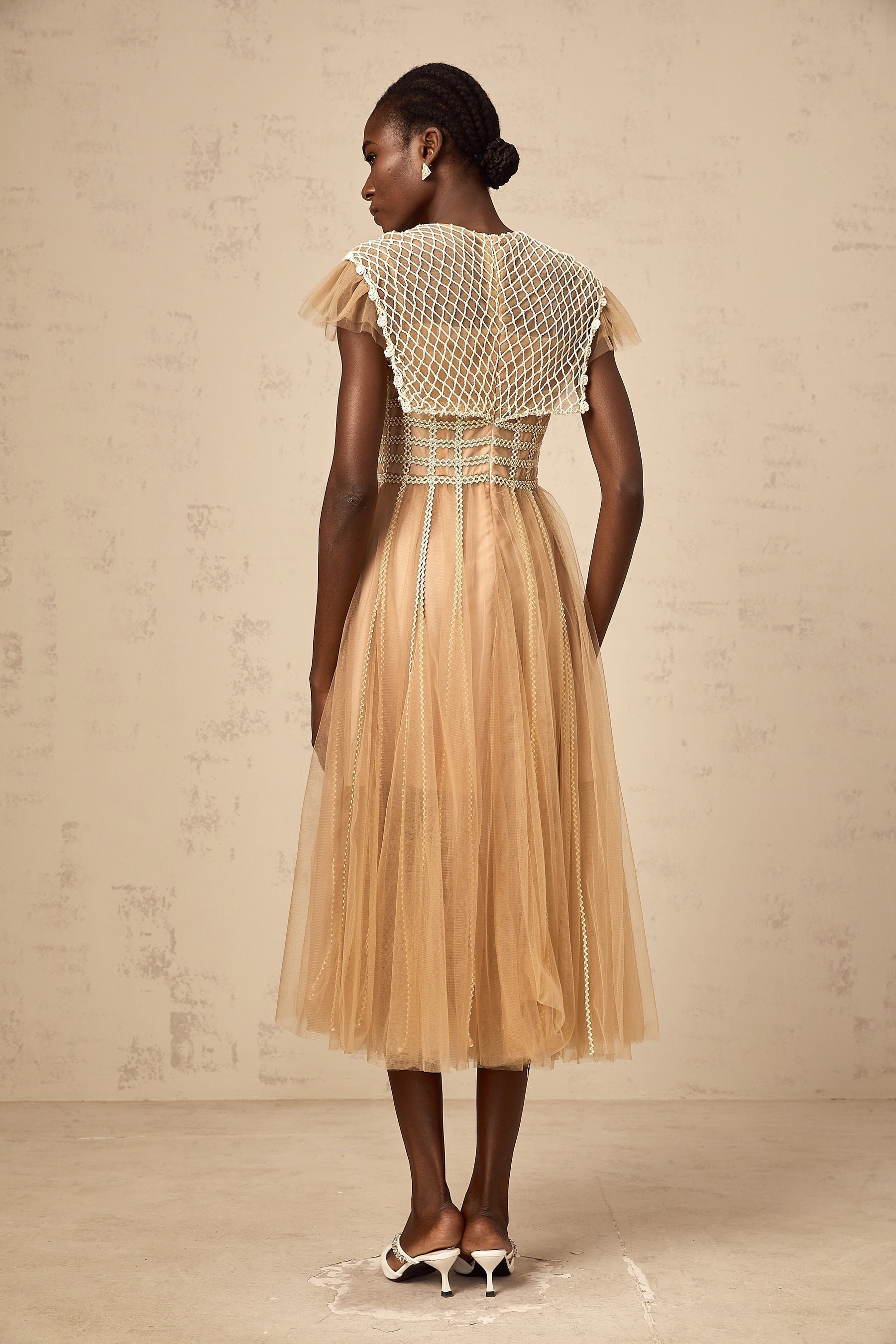 A woman wearing a neutralcolored midi dress with lace panels and tulle detailing standing in a relaxed pose