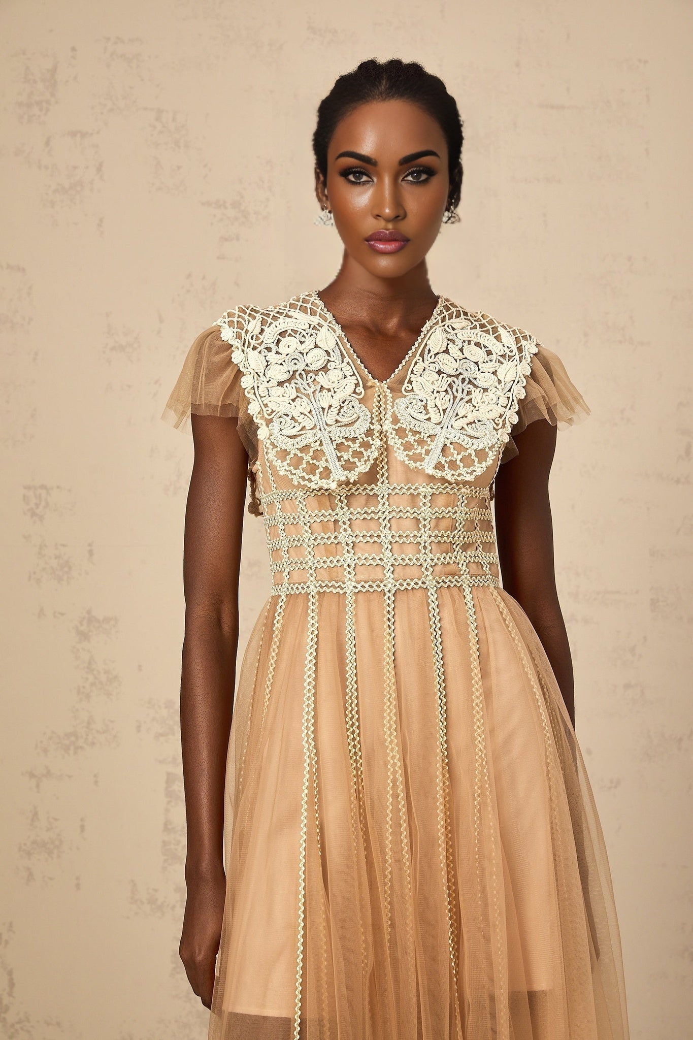A woman wearing a neutralcolored midi dress with lace panels and tulle detailing standing in a relaxed pose