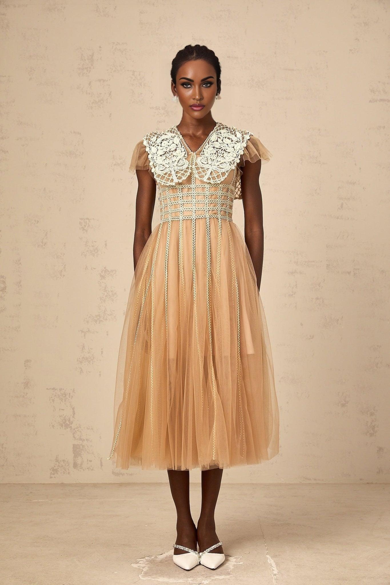 A woman wearing a neutralcolored midi dress with lace panels and tulle details standing in a relaxed pose