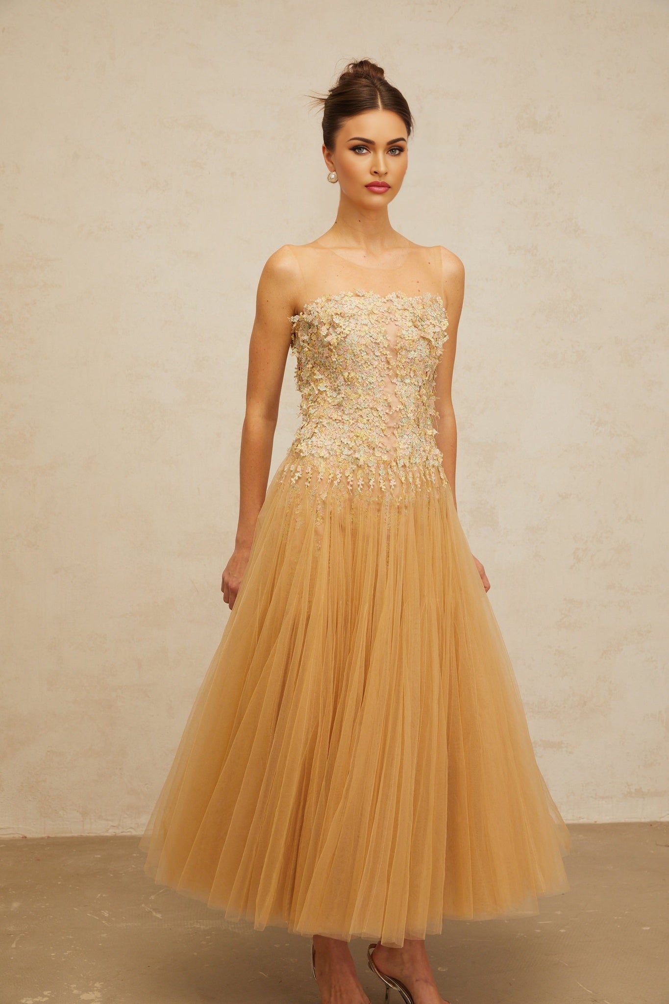 A woman wearing a neutralcolored midi dress with pleated tulle and sequin embellishments standing in an elegant pose