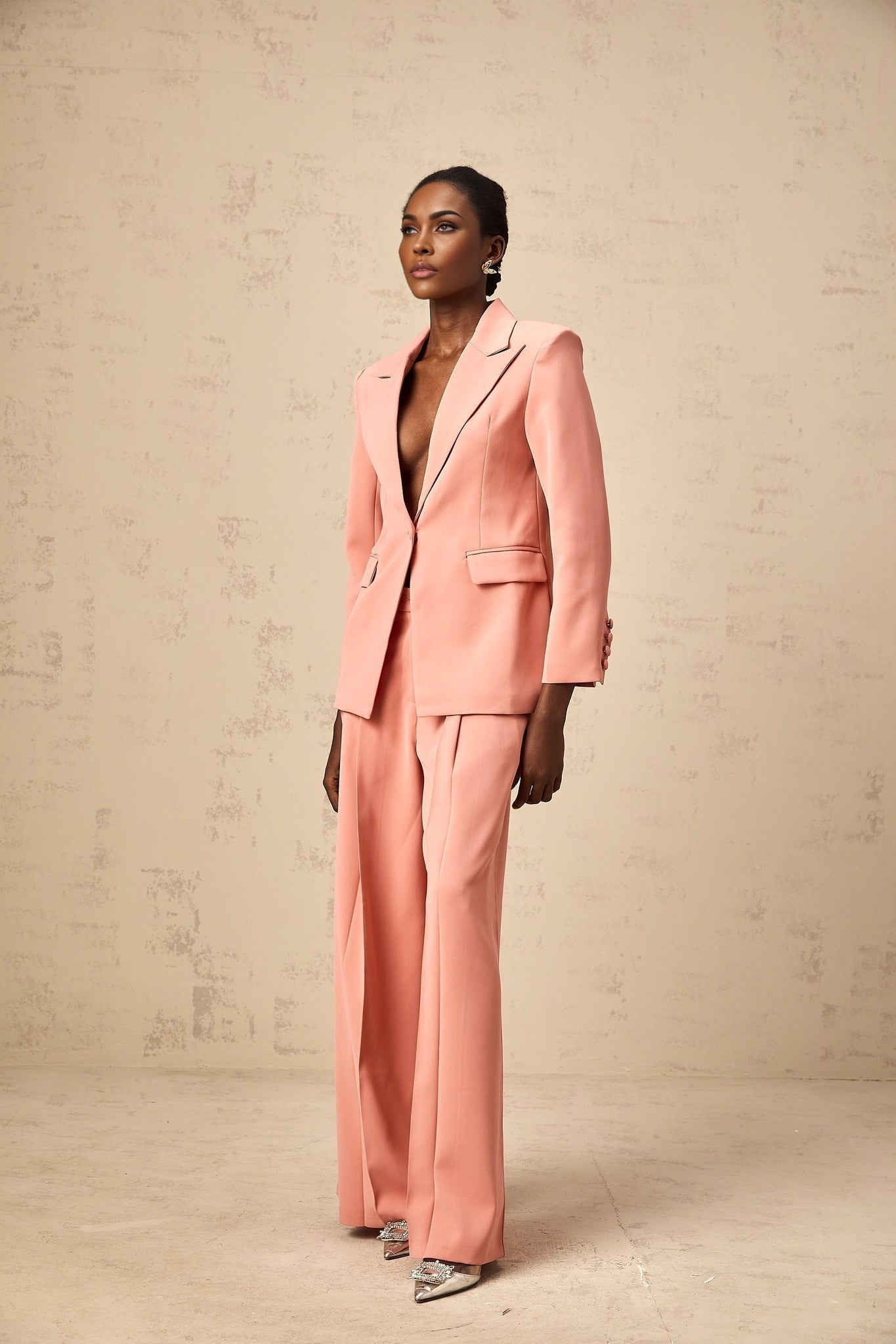 A woman wearing a pink blazer and trousers set with embroidery details and cutout designs The blazer features a tailored fit while the trousers are slim The set is adorned with delicate floral embroidery in contrasting colors Cutout elements add a modern touch to the ensemble